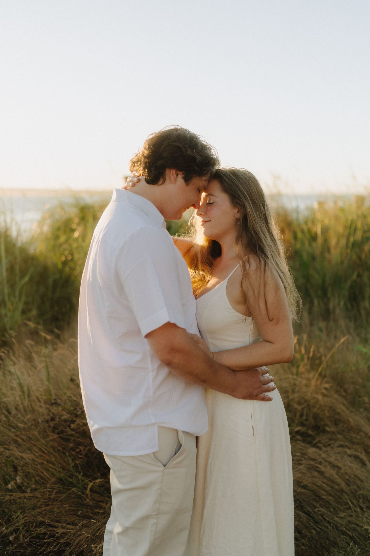 A romantic couple in white outfits embraces during a sunset photoshoot in a grassy field.