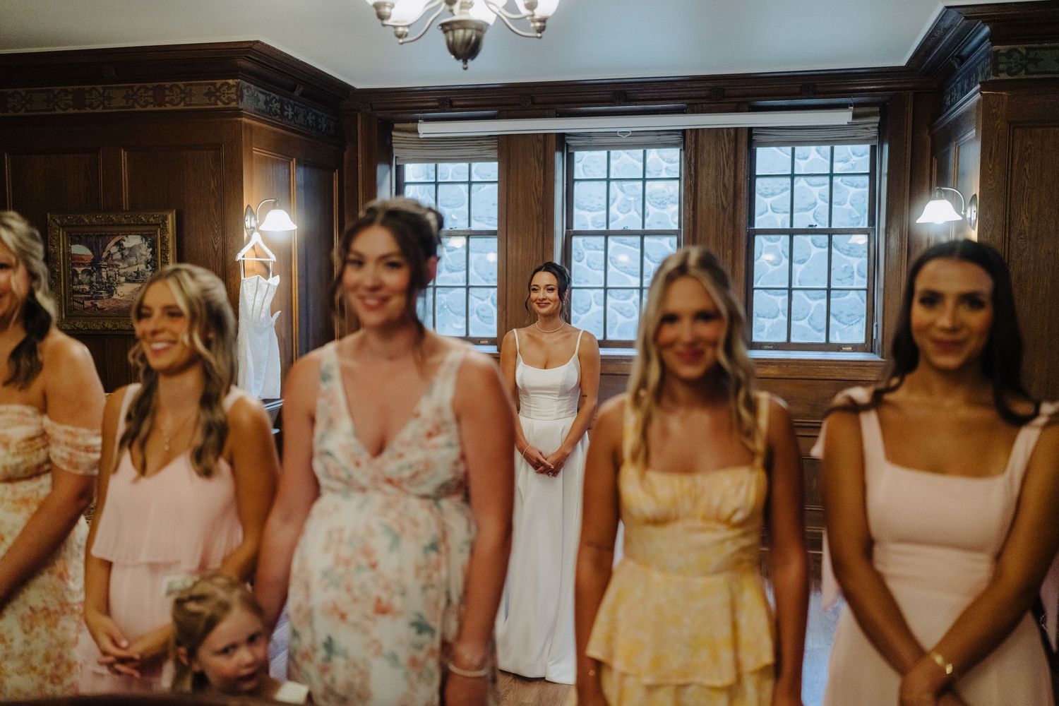 A group of bridesmaids wearing pastel dresses gather in a wood-paneled room before a wedding ceremony.