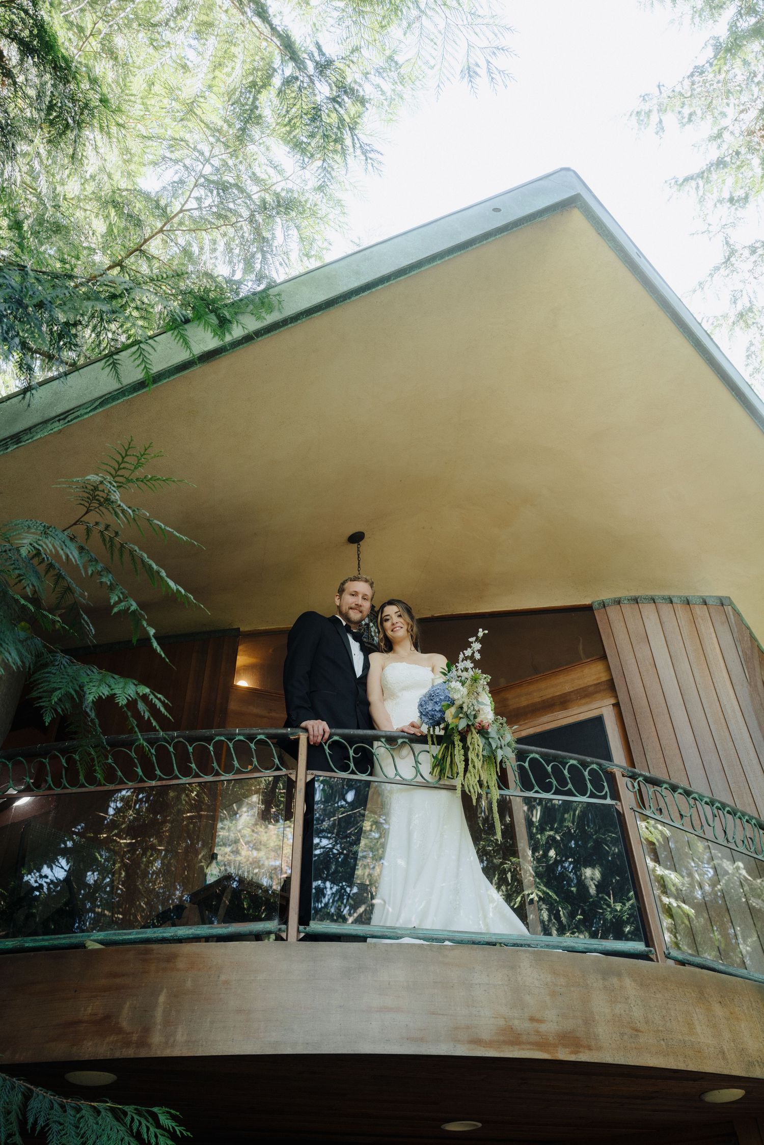 Modern architectural balcony with evergreen trees serves as backdrop for wedding photos.