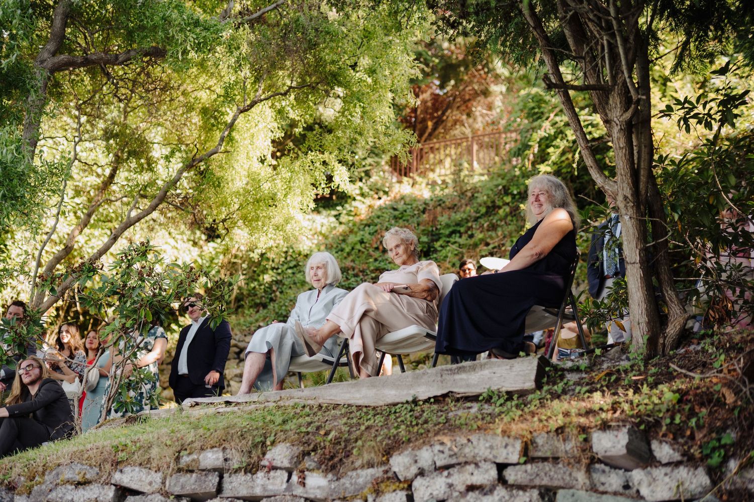 Wedding guests seated on stone walls in a lush garden setting during an outdoor celebration.