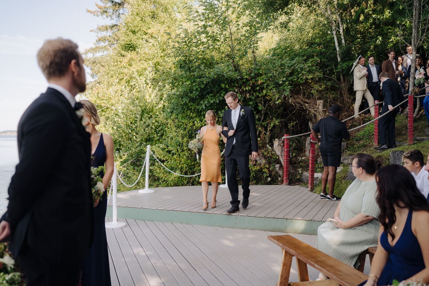 Wedding ceremony guests gather on a wooden deck surrounded by lush greenery during an outdoor celebration.