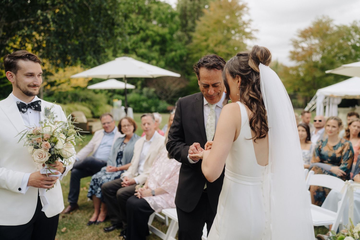 Outdoor garden wedding ceremony with guests seated on white chairs and floral decorations.