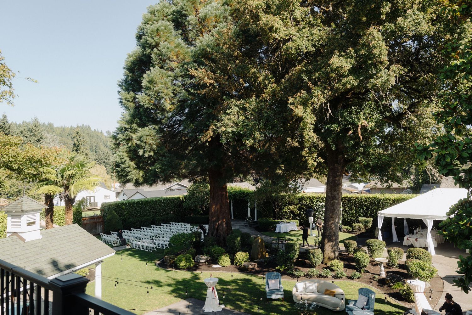 Aerial view of an outdoor wedding venue with white chairs and tables set up under large trees.