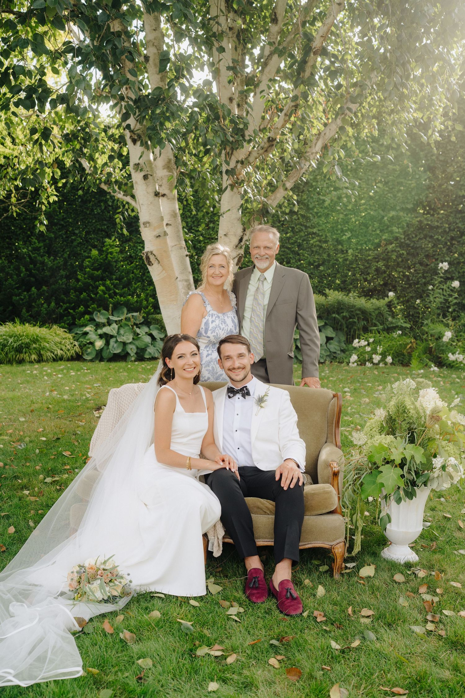 Wedding party poses on vintage armchair under birch trees in garden setting.