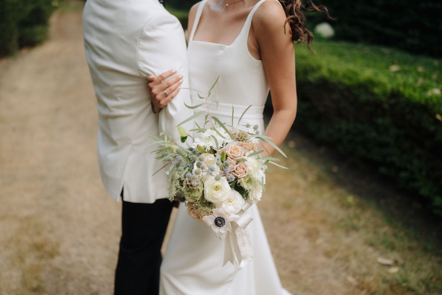 Close-up details of white wedding bouquet and dress against natural outdoor backdrop.