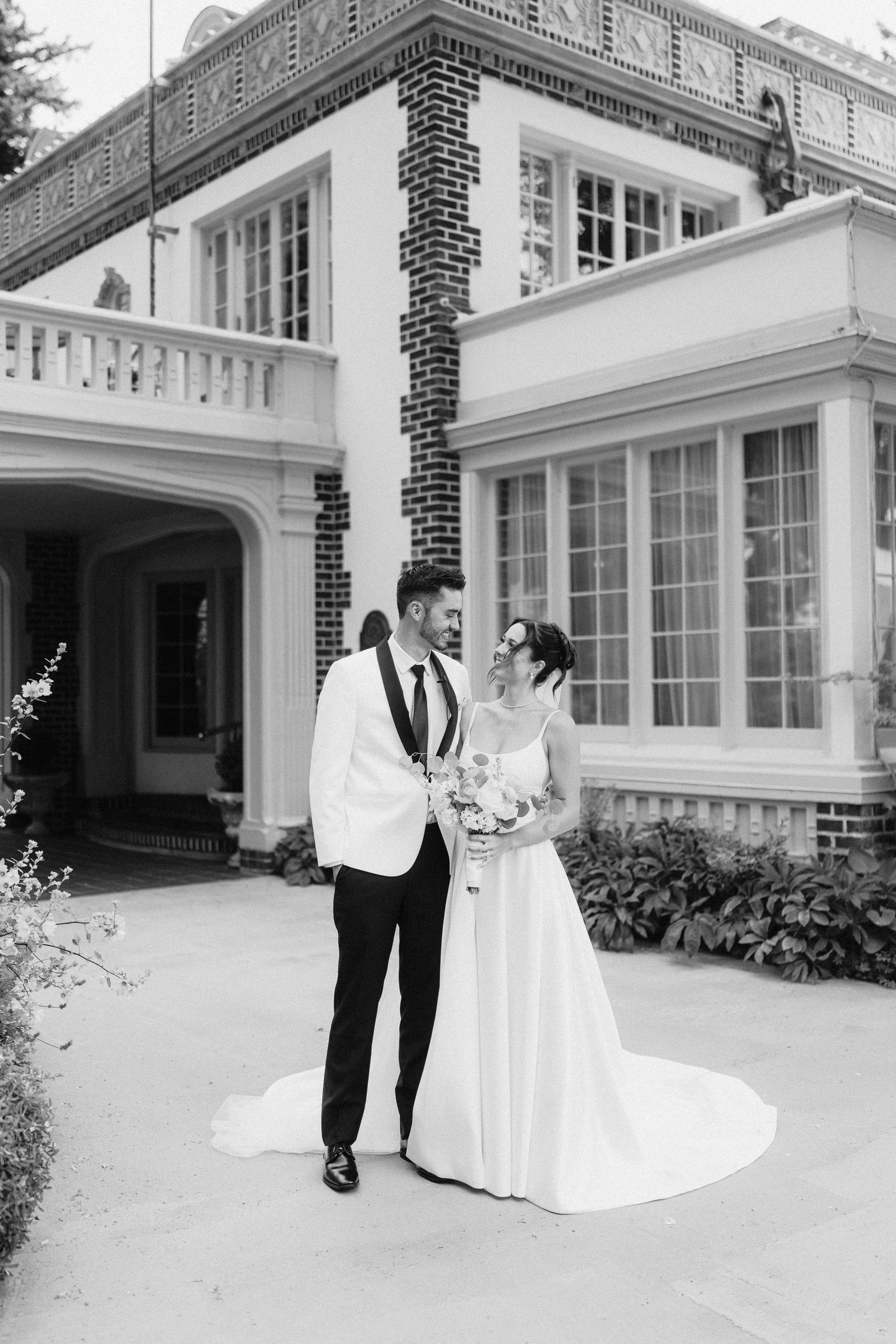 A wedding couple poses outside a grand white mansion with contrasting black brick details.