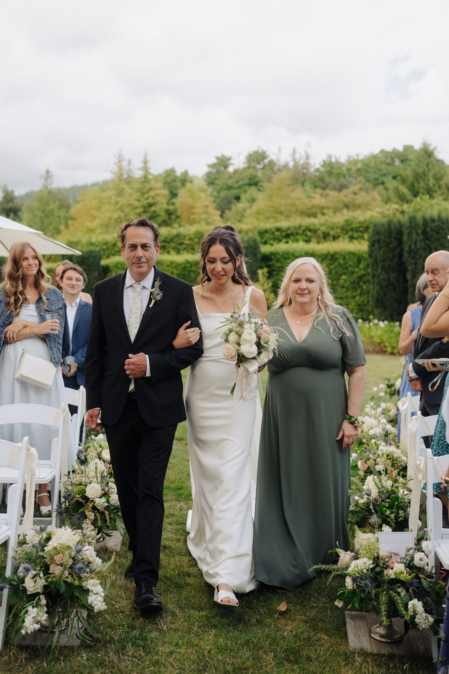 Wedding party walks down outdoor aisle decorated with white floral arrangements on a grassy lawn.