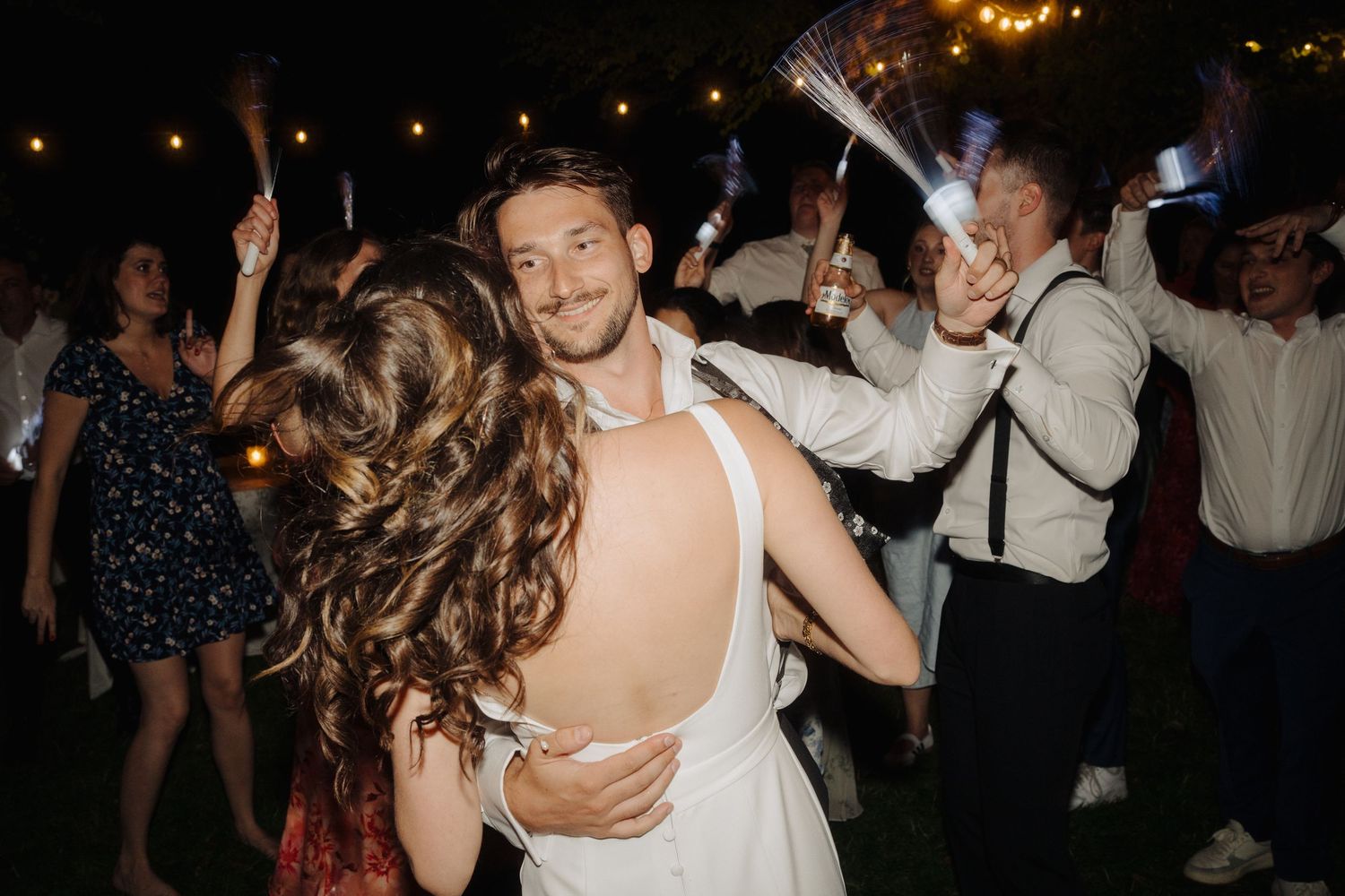 Wedding reception guests dance and celebrate under dim evening lighting.