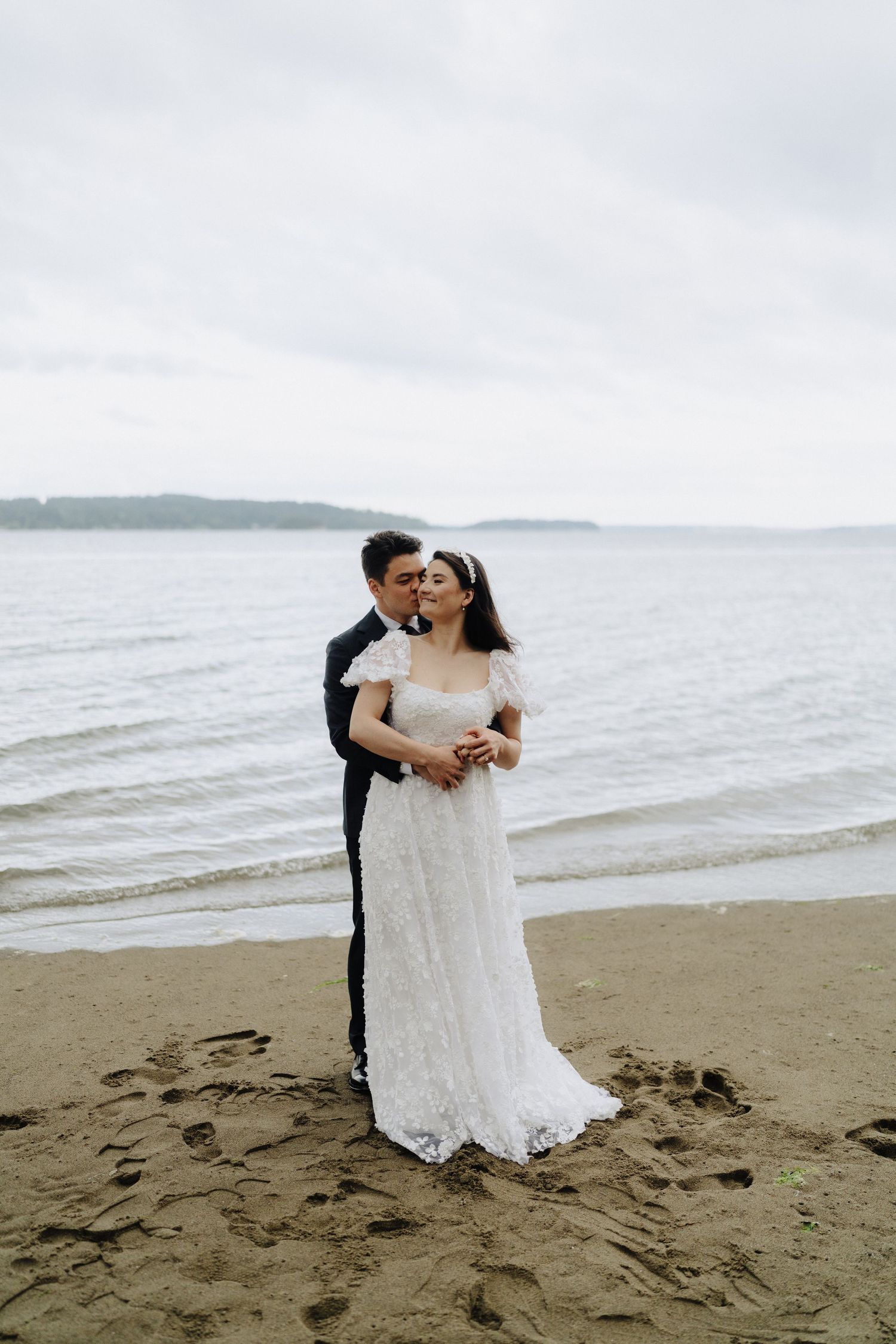 A couple shares an intimate moment on a cloudy beach during their wedding photoshoot.