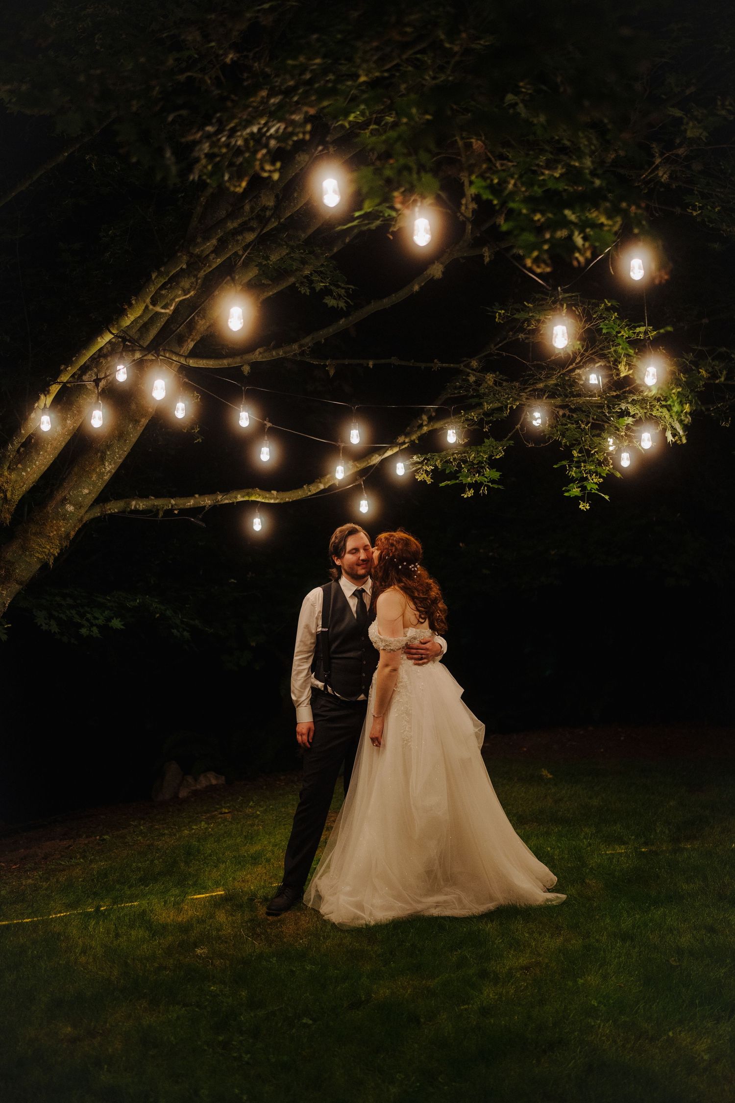 Bride and groom sharing romantic moment under glowing string lights in a lush garden at night.
