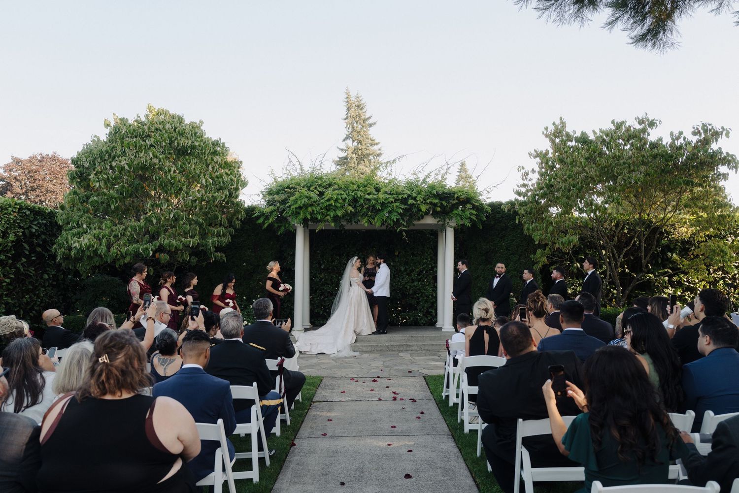 Outdoor wedding ceremony with guests seated facing a white pergola.