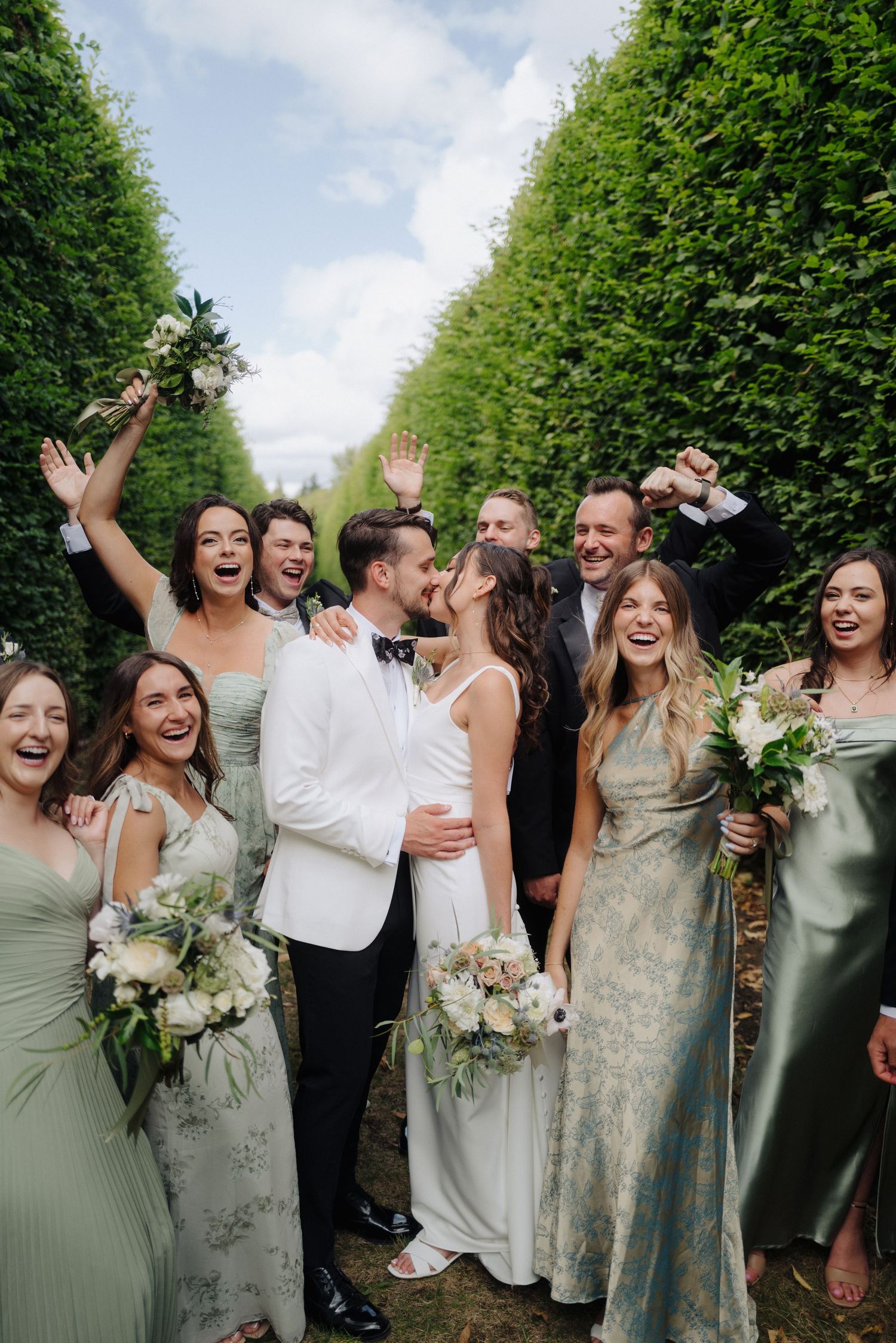 Wedding party celebrating with raised arms and joyful expressions in front of a hedge wall.