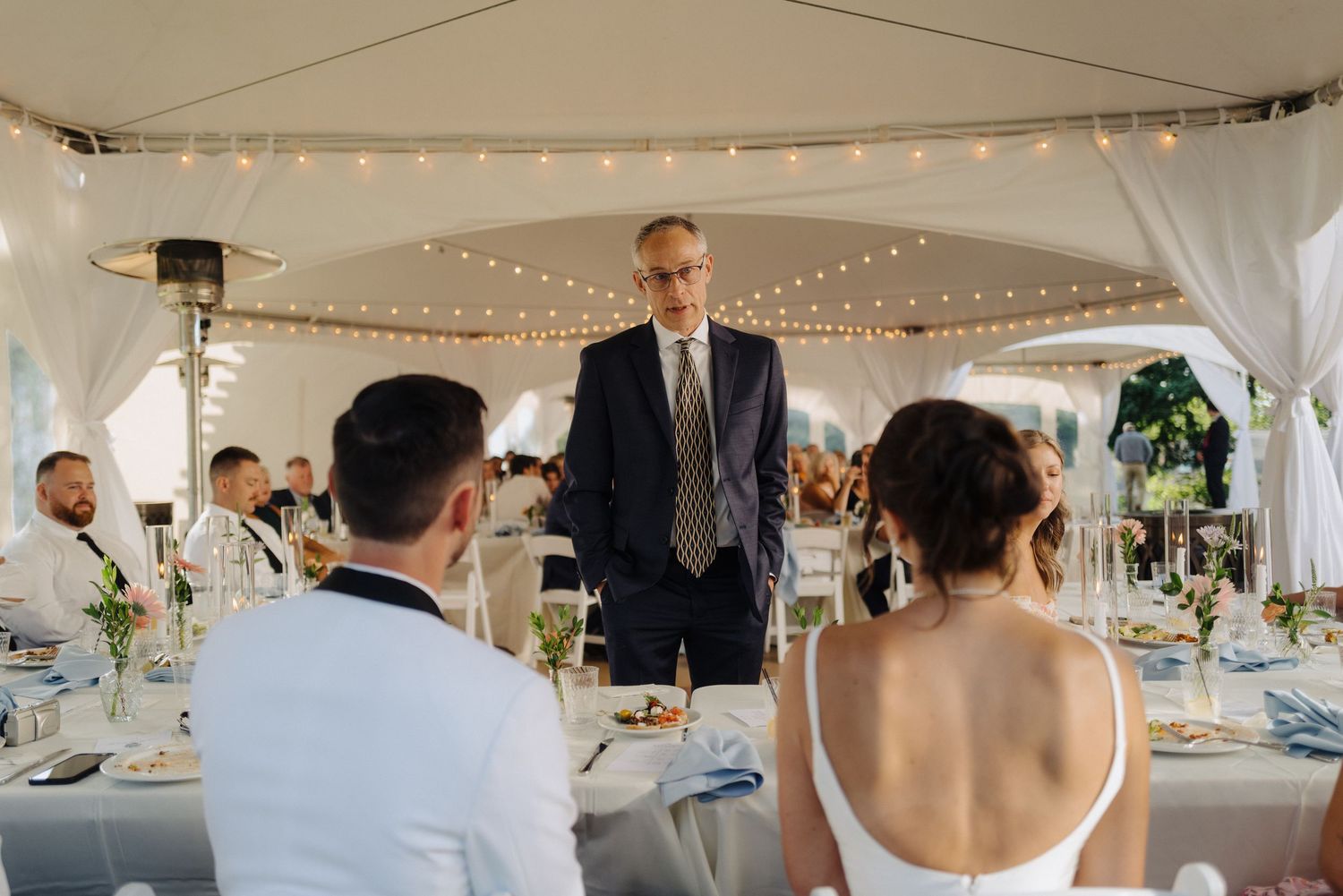 Wedding reception setup under white tent with string lights and elegant table settings.