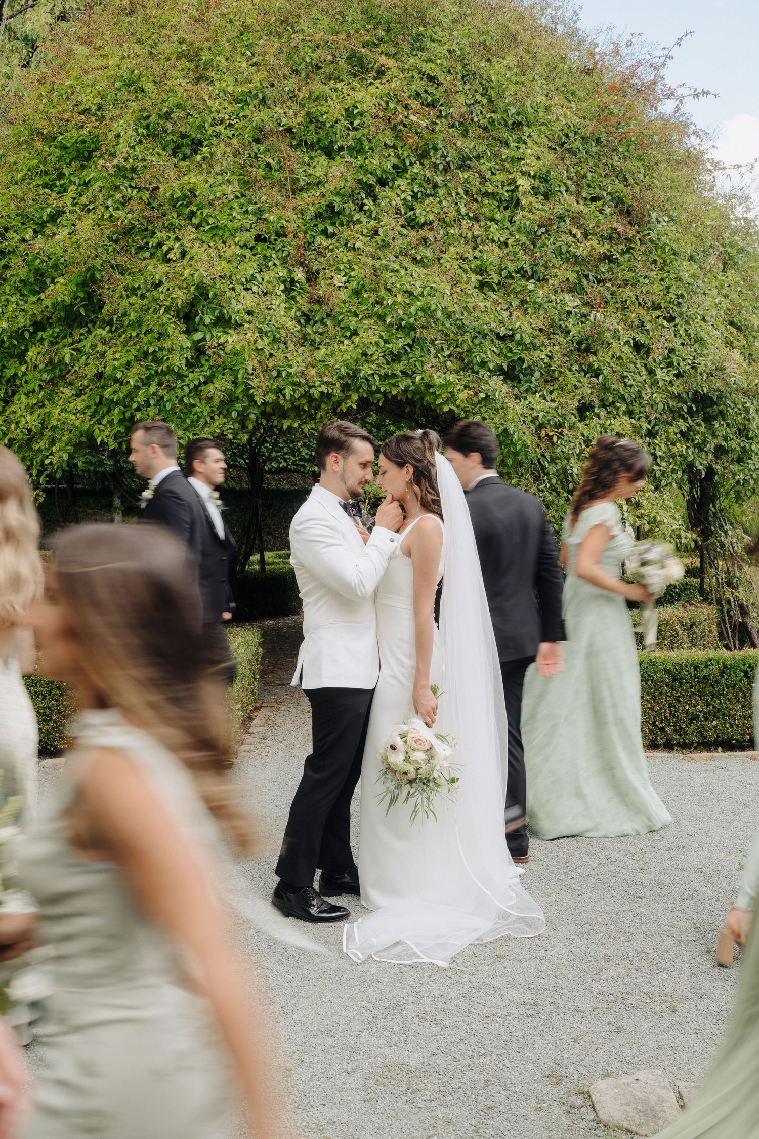 Wedding party in sage green dresses and formal attire gather for outdoor photos.