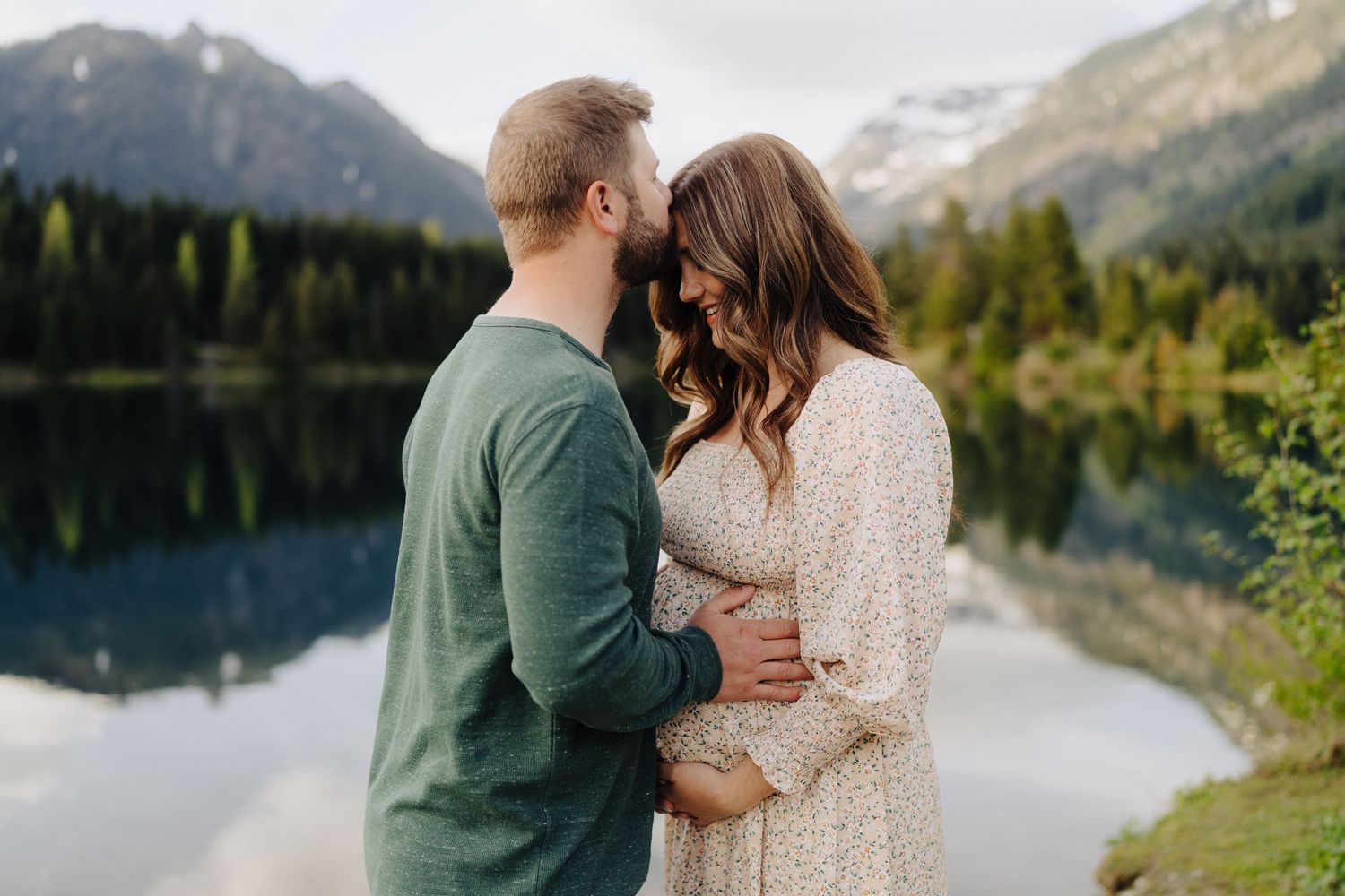 Romantic couple embraces near a scenic mountain lake with evergreen trees and peaks in the background.