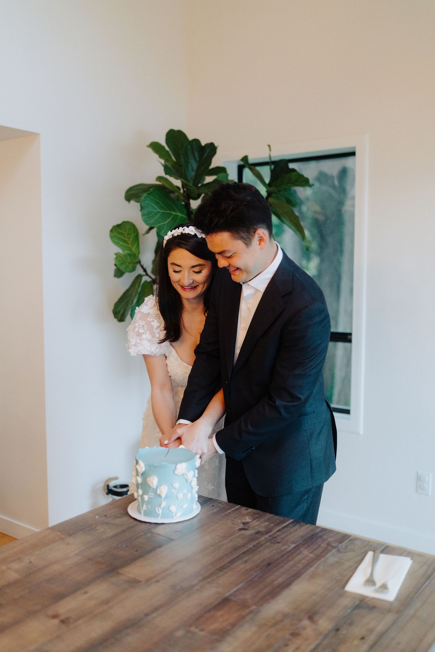 A newlywed couple leans over a light blue wedding cake on a wooden table with indoor plants in background.