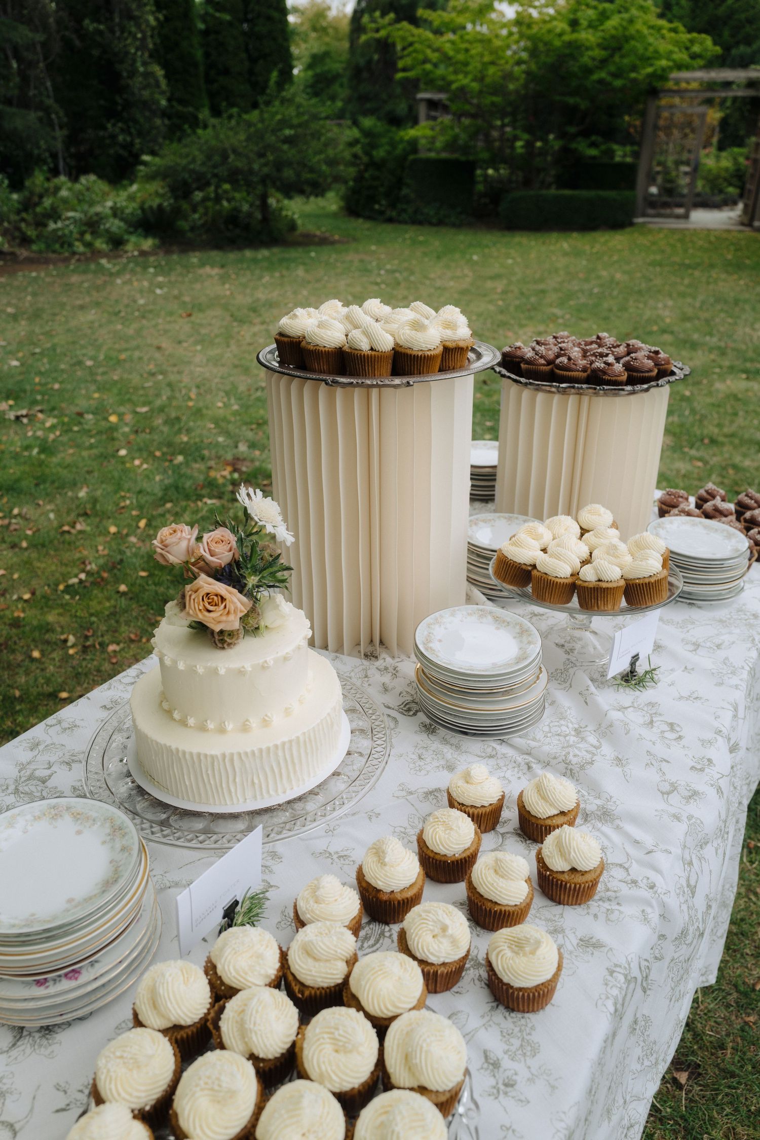 A wedding dessert table displays white frosted cupcakes and a tiered cake on lace tablecloths.