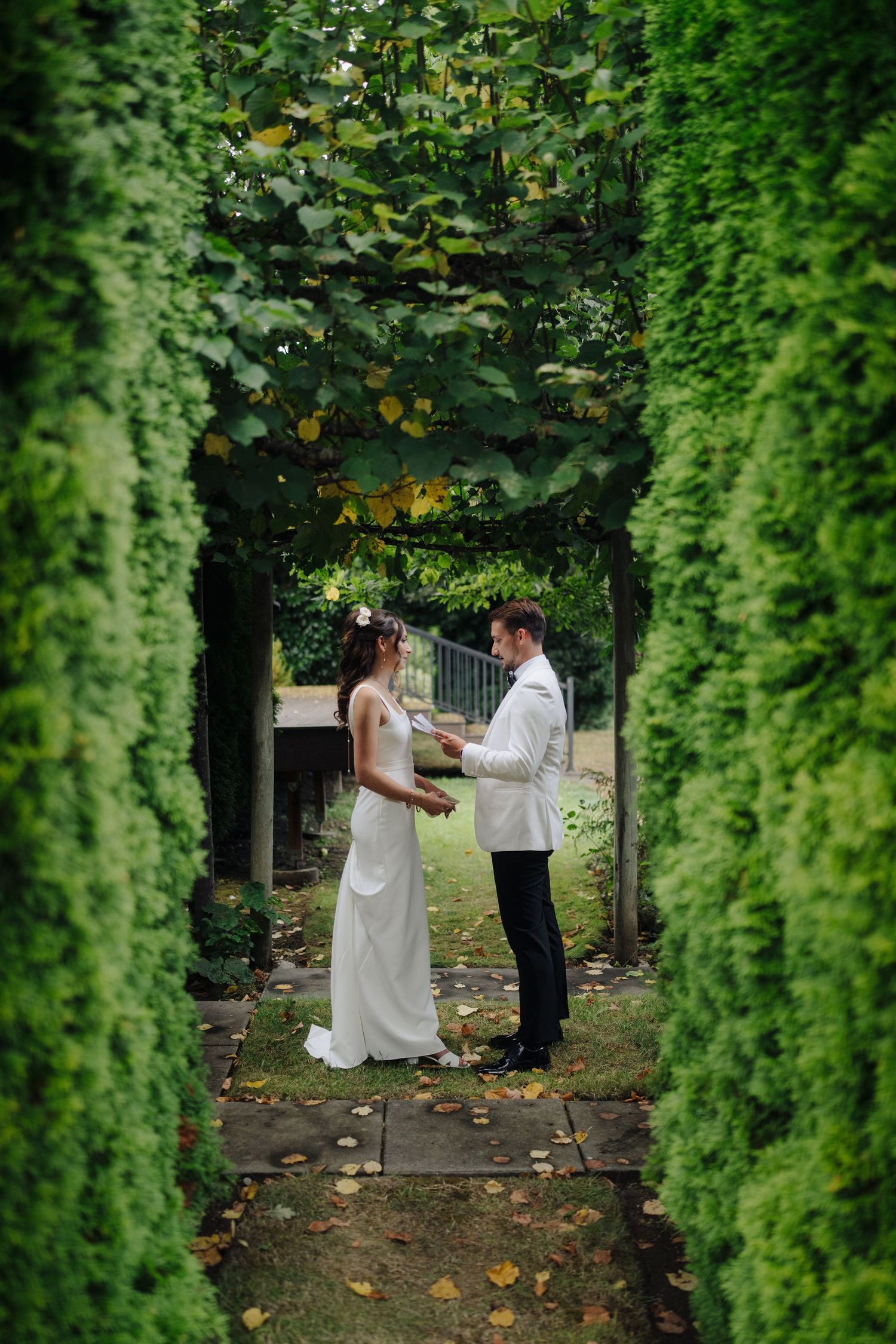 A couple shares a romantic moment on a tree-lined garden path with autumn leaves falling around them.