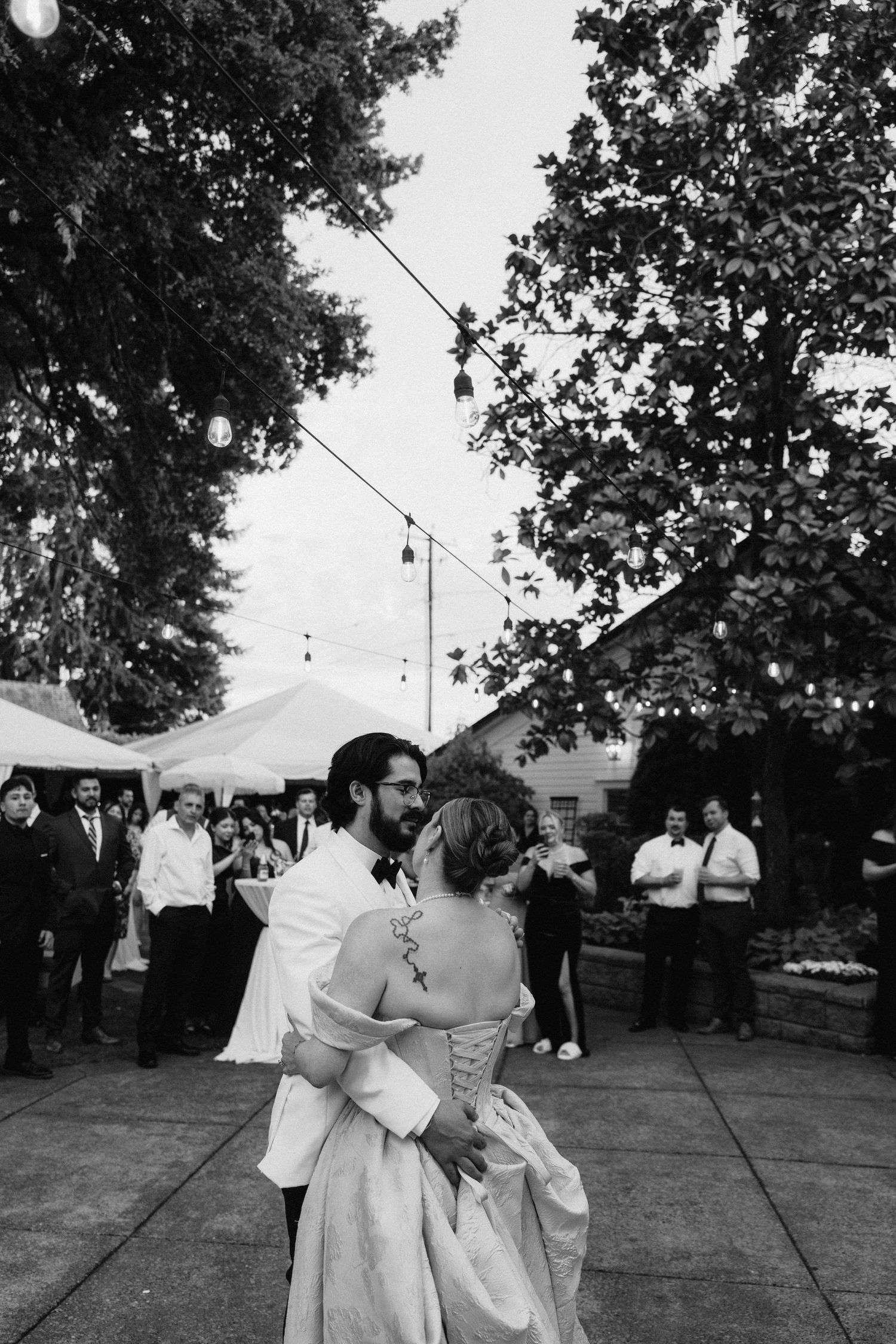 Black and white photo of couples dancing under string lights at outdoor wedding reception.