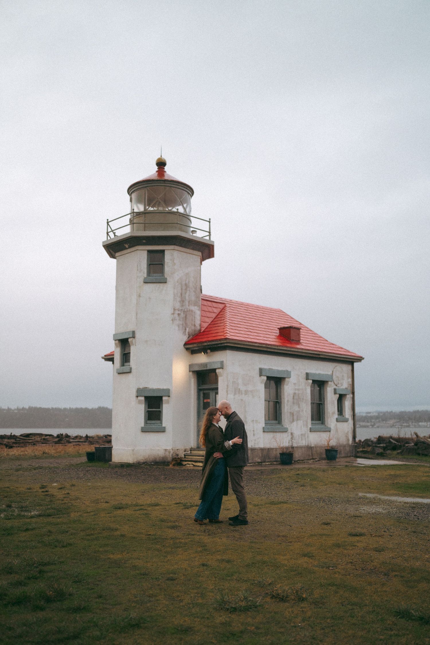 Couple sharing a tender moment in front of a historic white lighthouse with red roof.