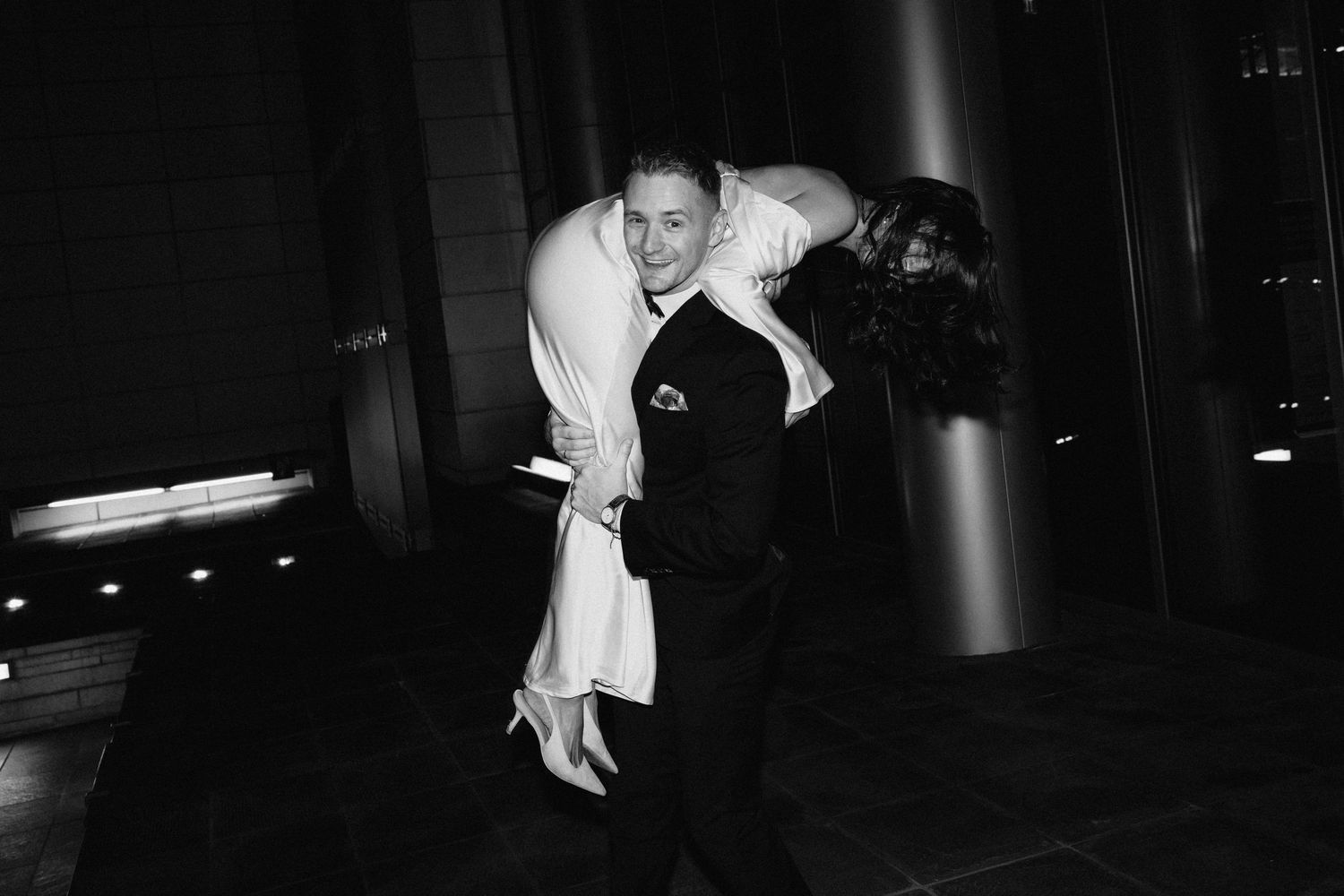 Groom joyfully lifts bride in elegant black and white nighttime wedding celebration photo.