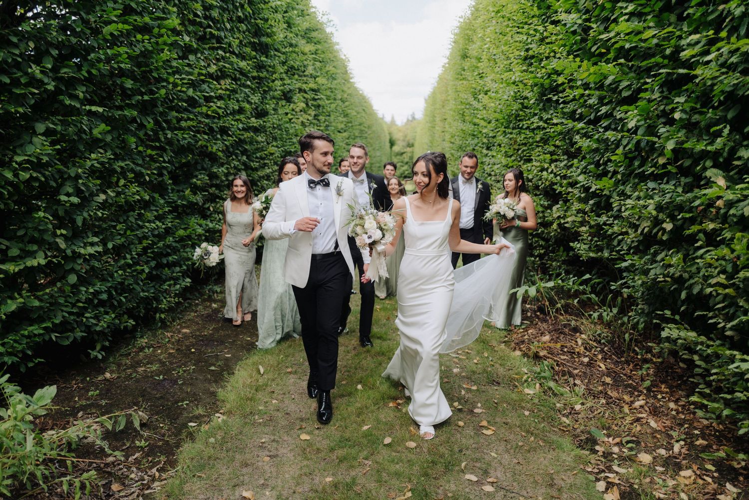Wedding party walking together down a green hedge-lined pathway.