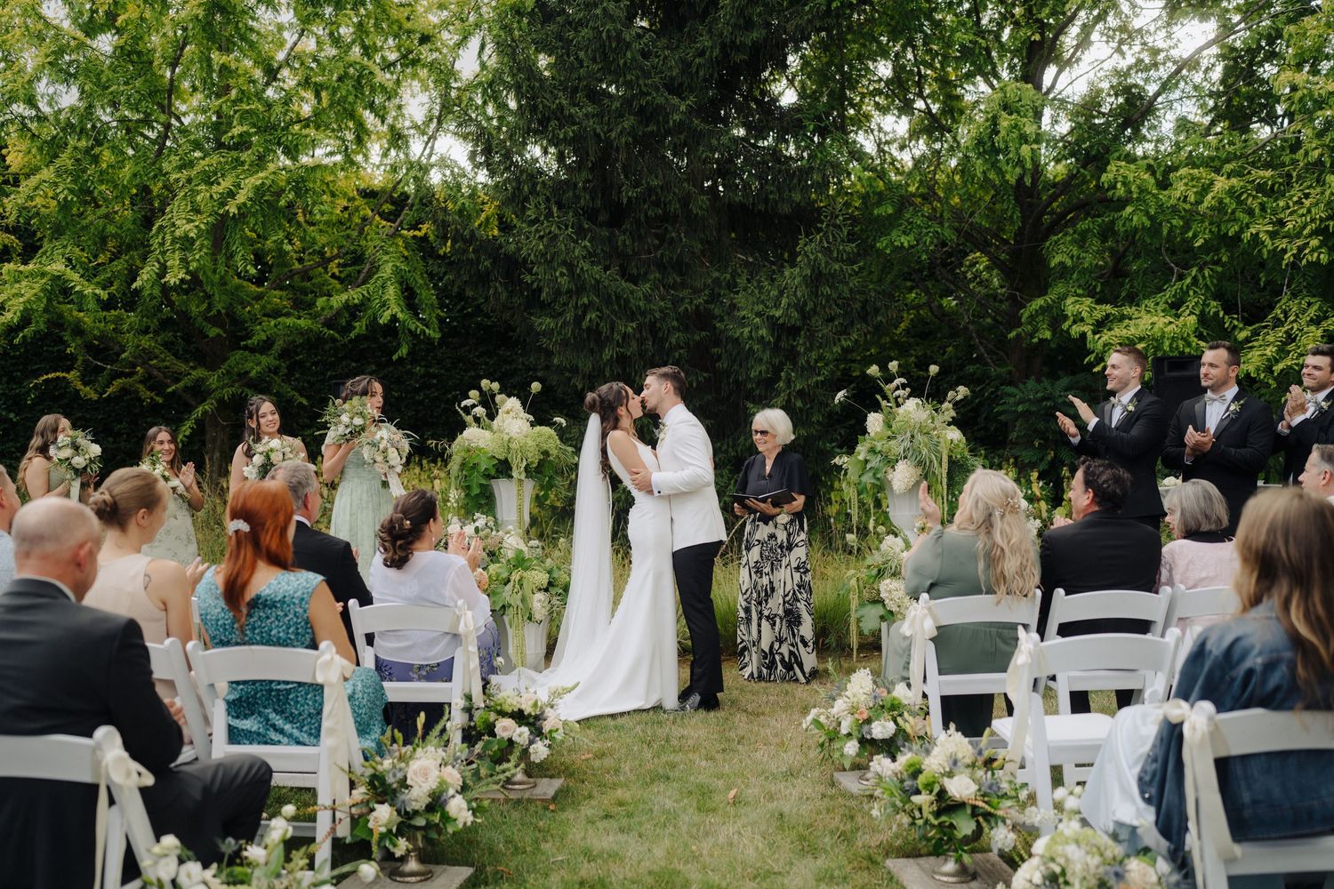 Wedding guests seated in white chairs on a lawn during an outdoor ceremony with natural greenery backdrop.
