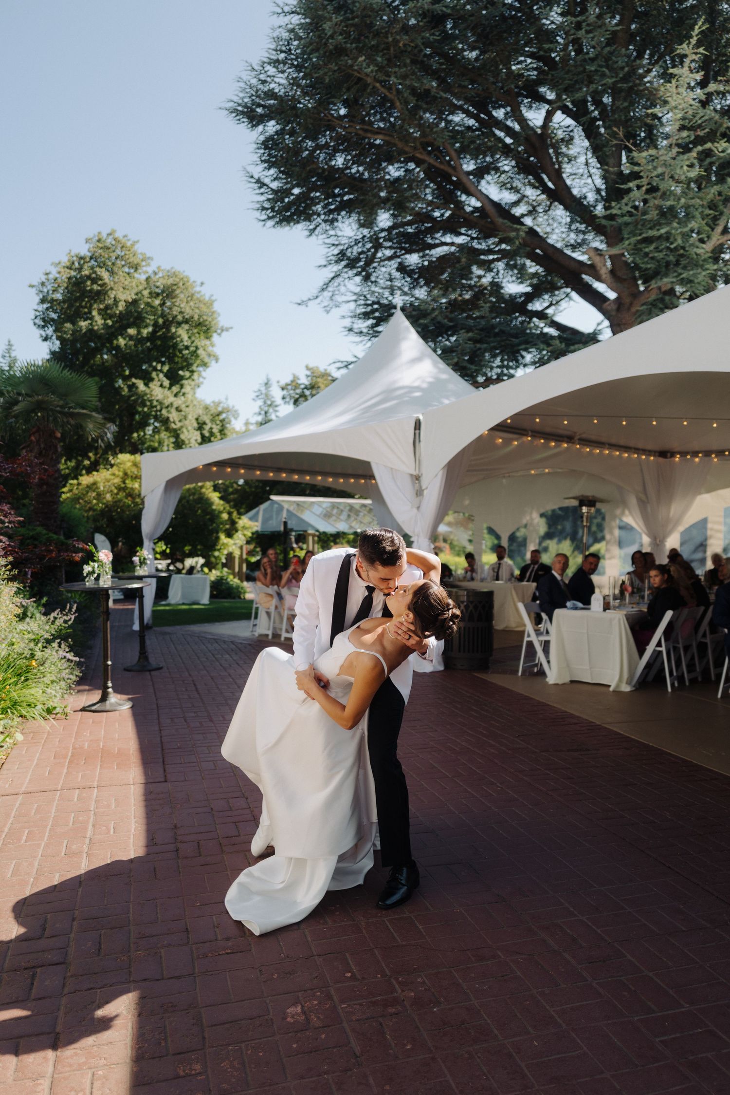 Wedding couple dancing under outdoor tent with romantic lighting and guests watching.