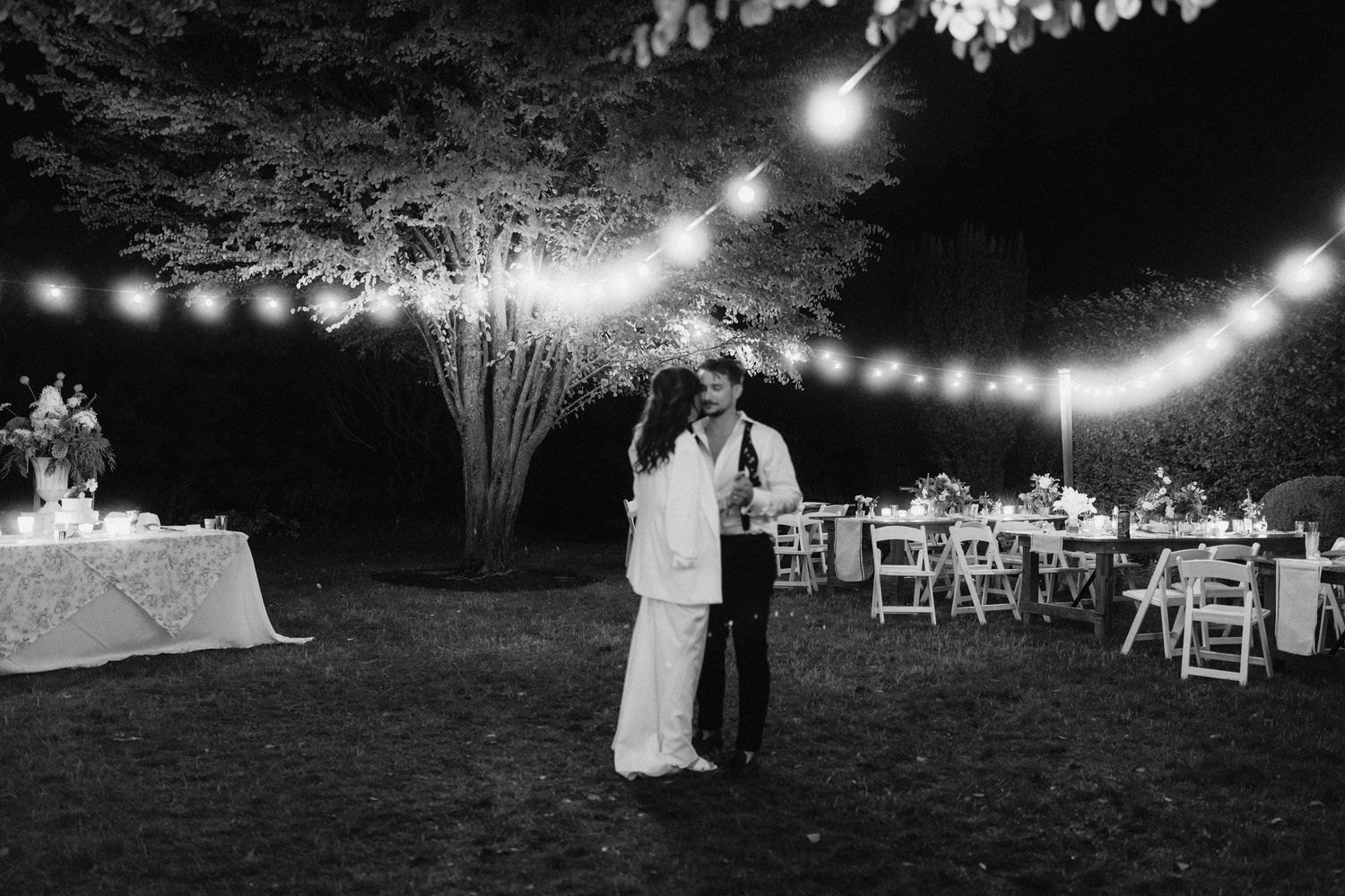 String lights illuminate an outdoor wedding reception setup with tables and chairs at dusk.