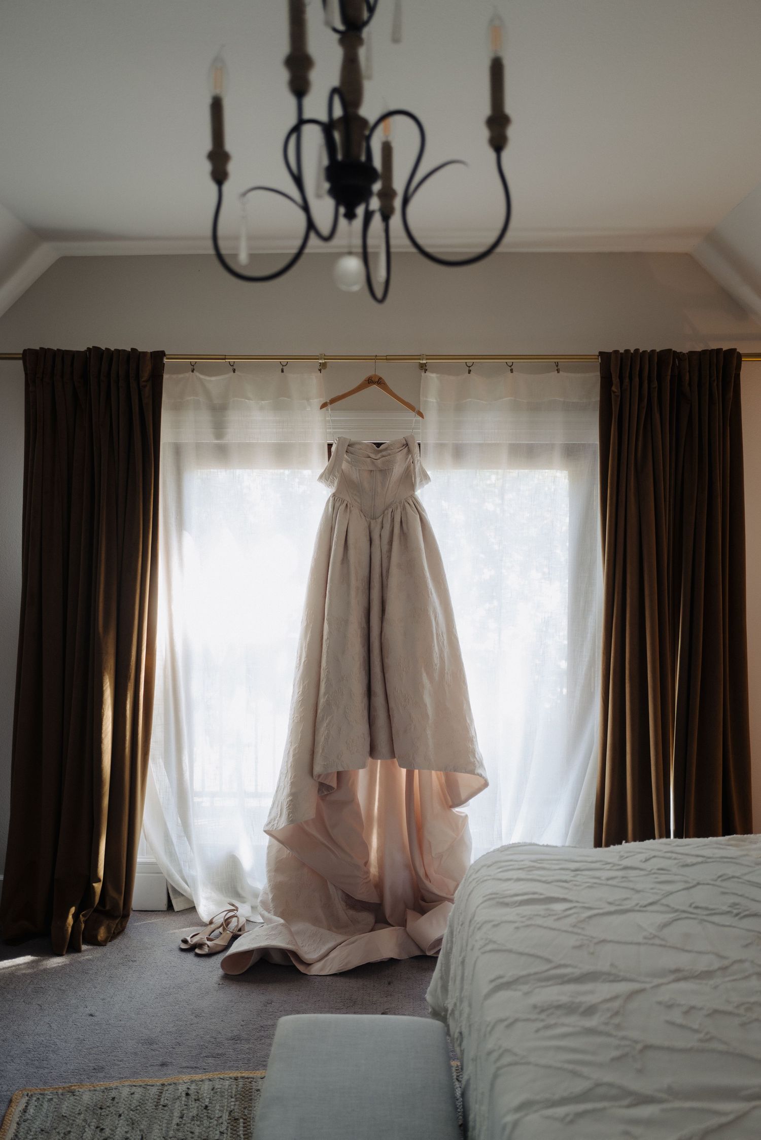 An elegant white wedding dress hangs by a window with dark curtains beneath a vintage chandelier.
