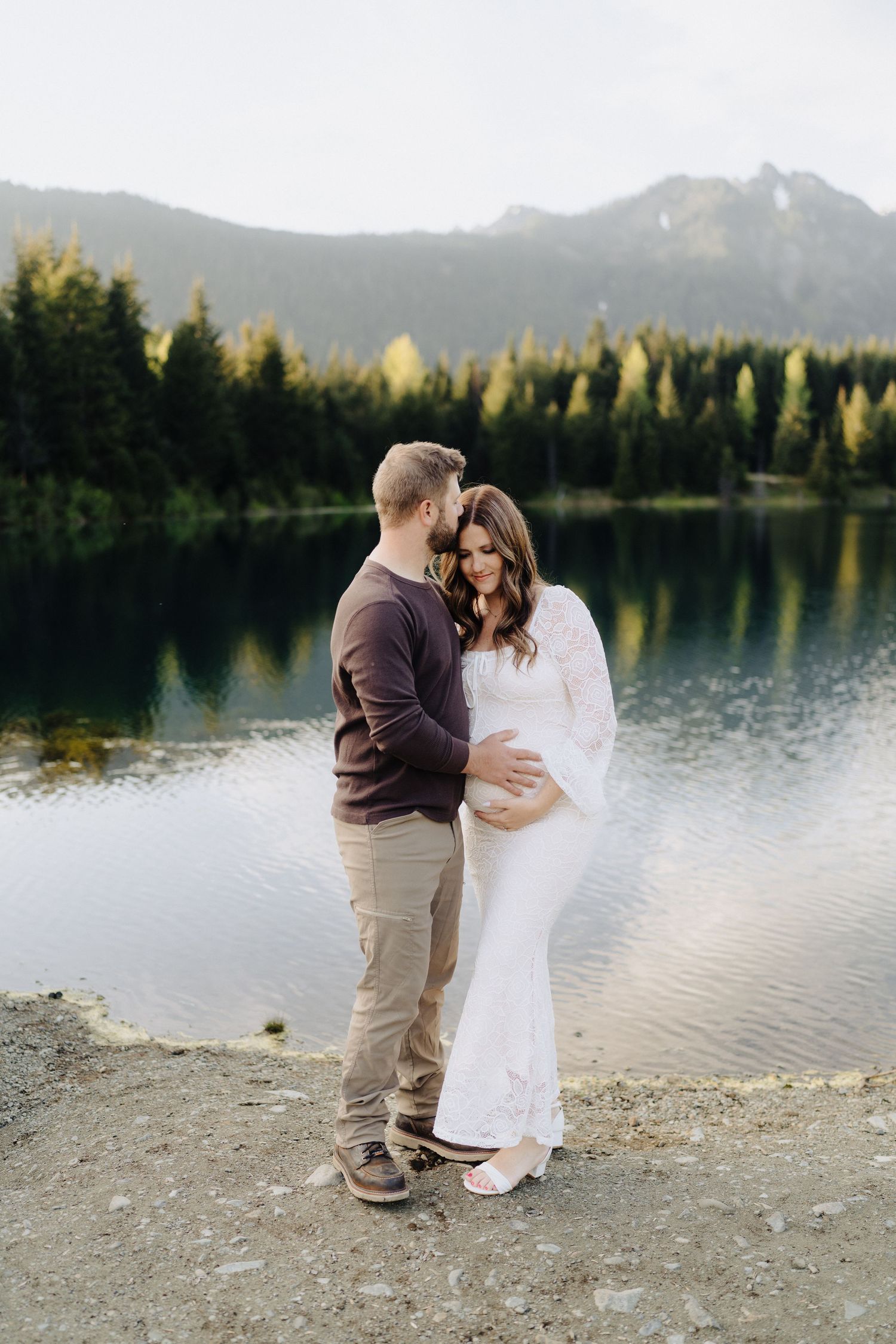 A romantic couple embraces by a serene mountain lake during golden hour with evergreen trees in the background.
