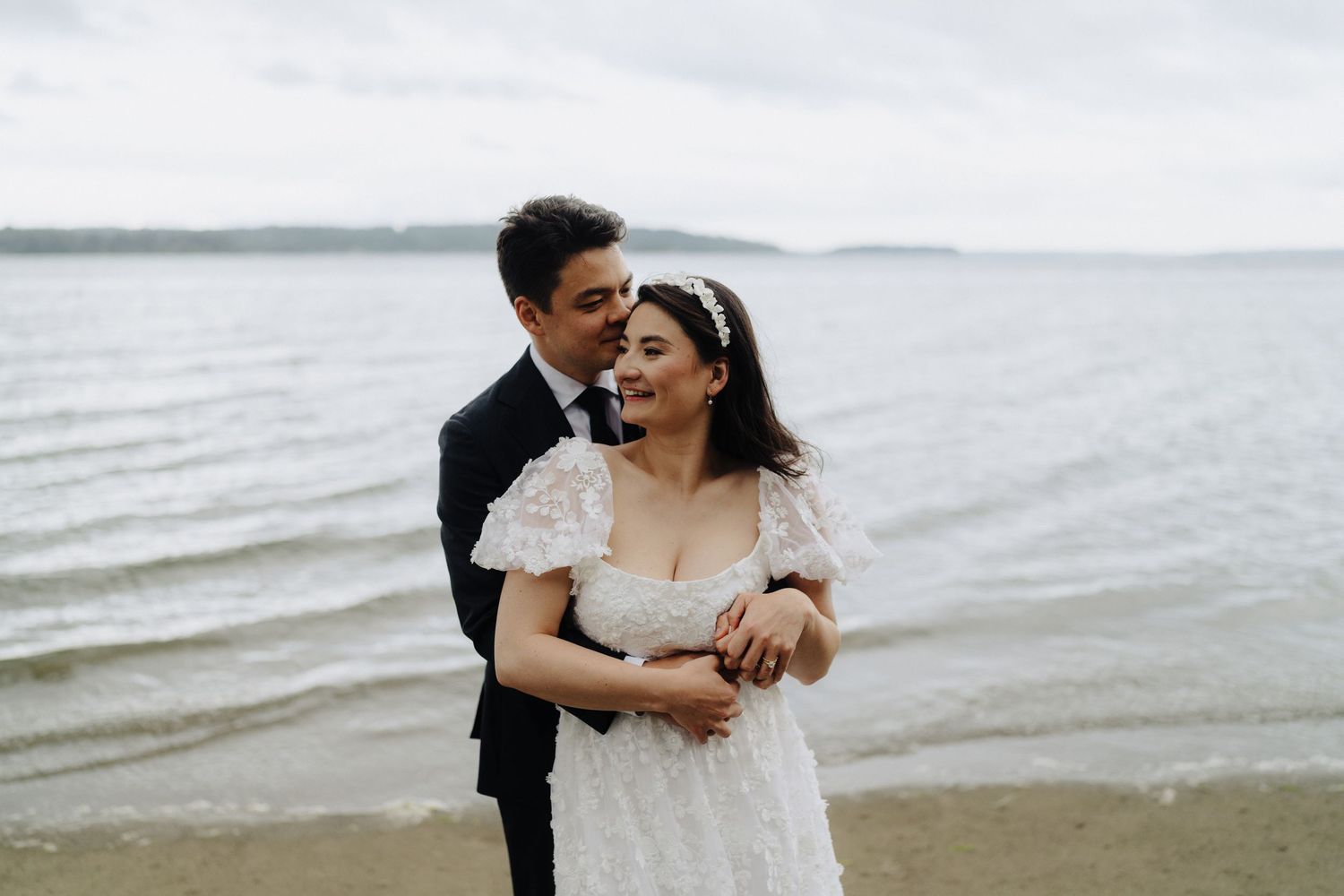 Newlyweds embrace on a cloudy beach with waves in the background.