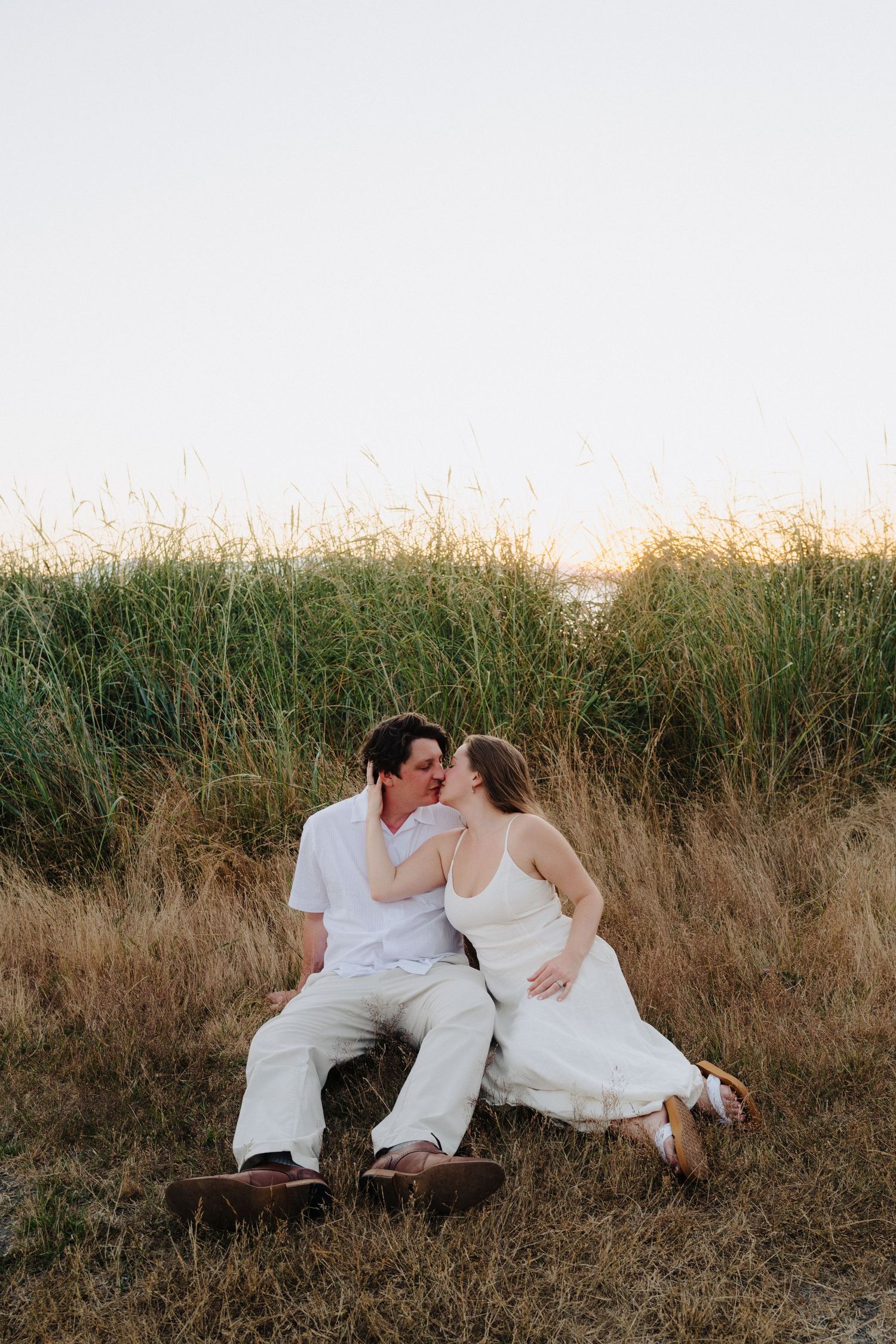 Couple in white clothing lying together in tall grass during a sunset photoshoot.