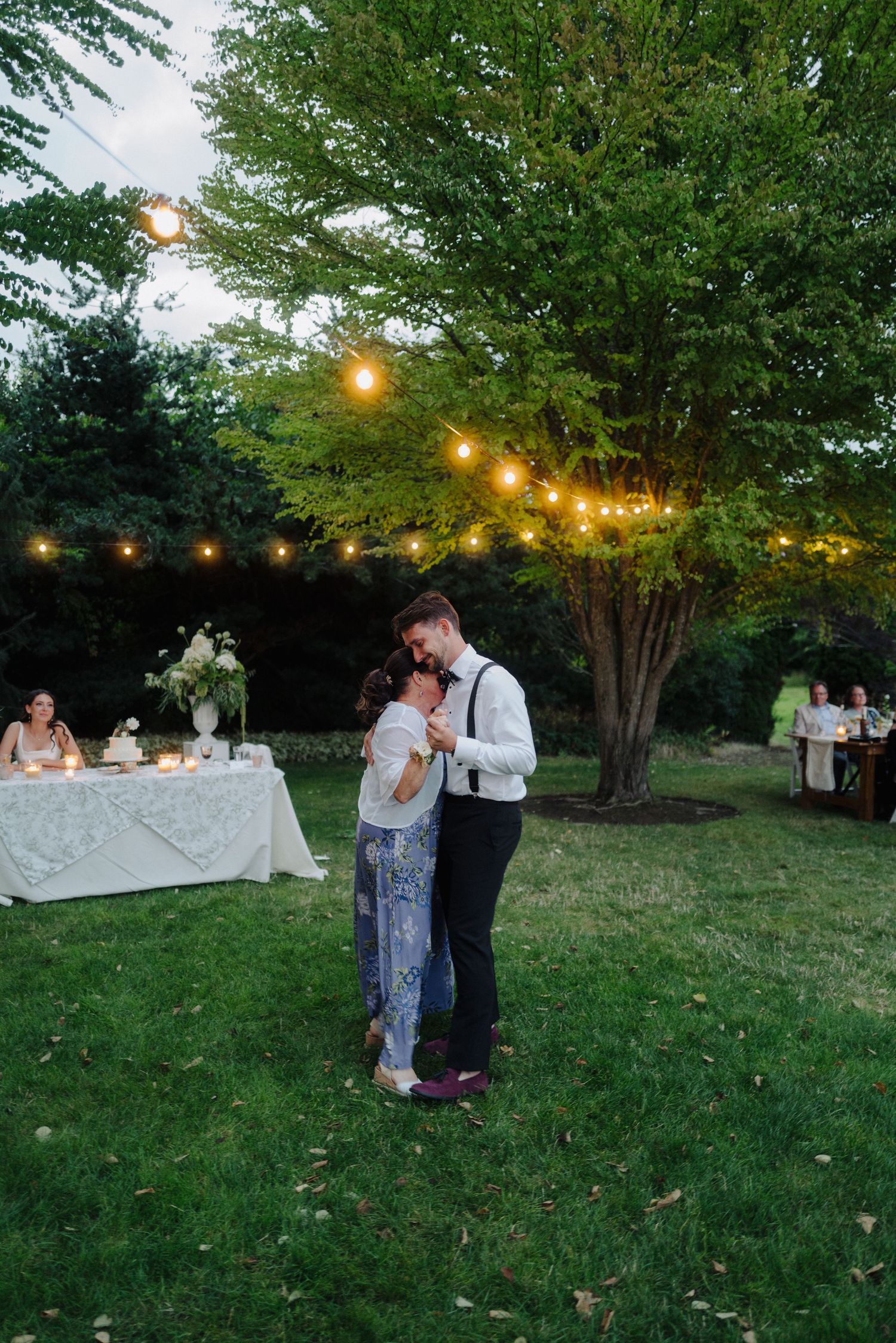 Wedding couple dances at twilight under string lights in garden setting.