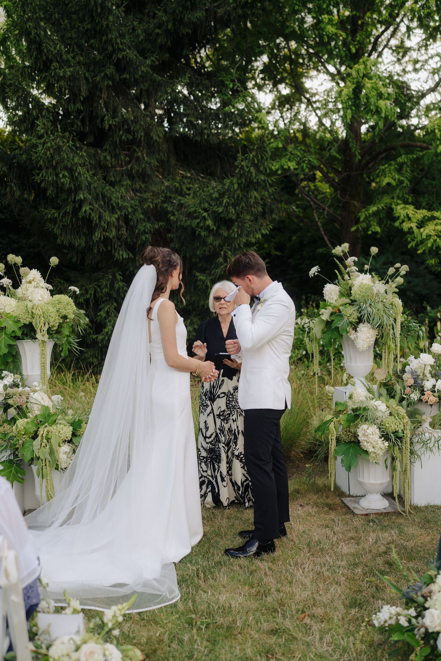 Outdoor wedding ceremony in progress with white floral decorations and natural garden setting.