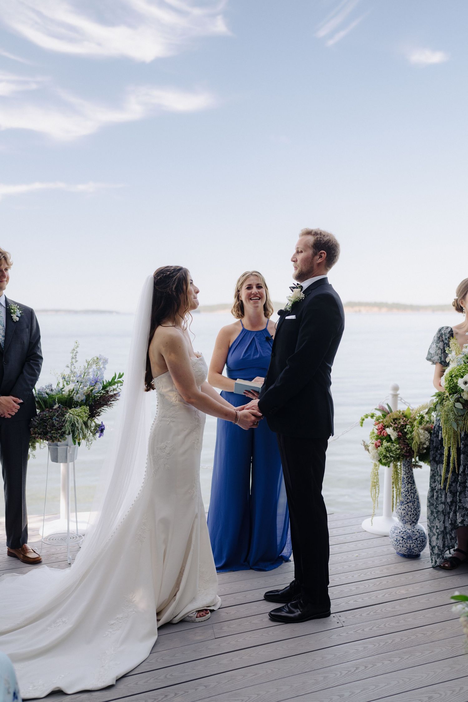 A wedding ceremony takes place on a scenic waterfront deck with guests gathered around the couple and officiant.