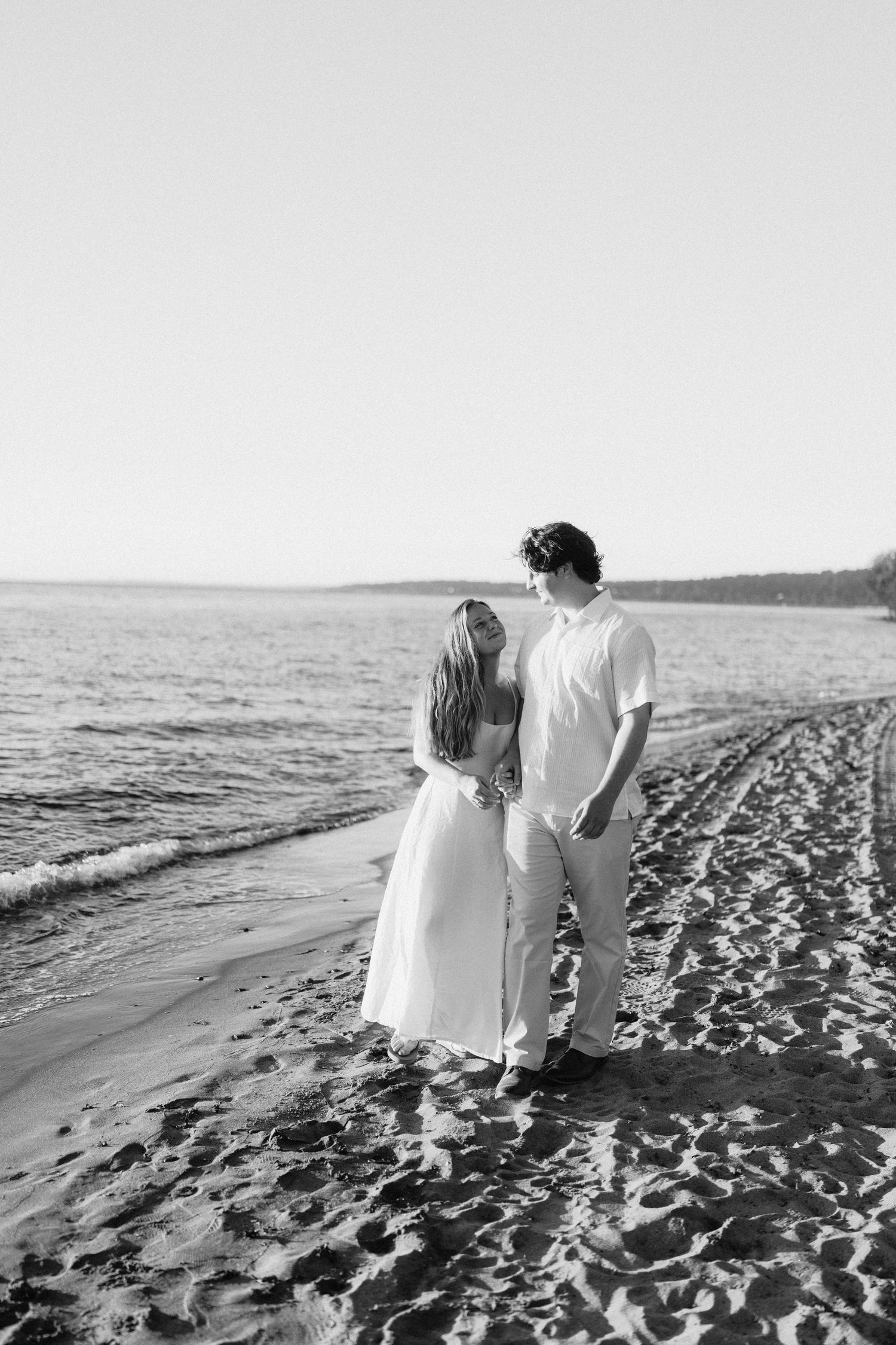 A couple in white outfits walks hand in hand along a sandy beach shoreline.