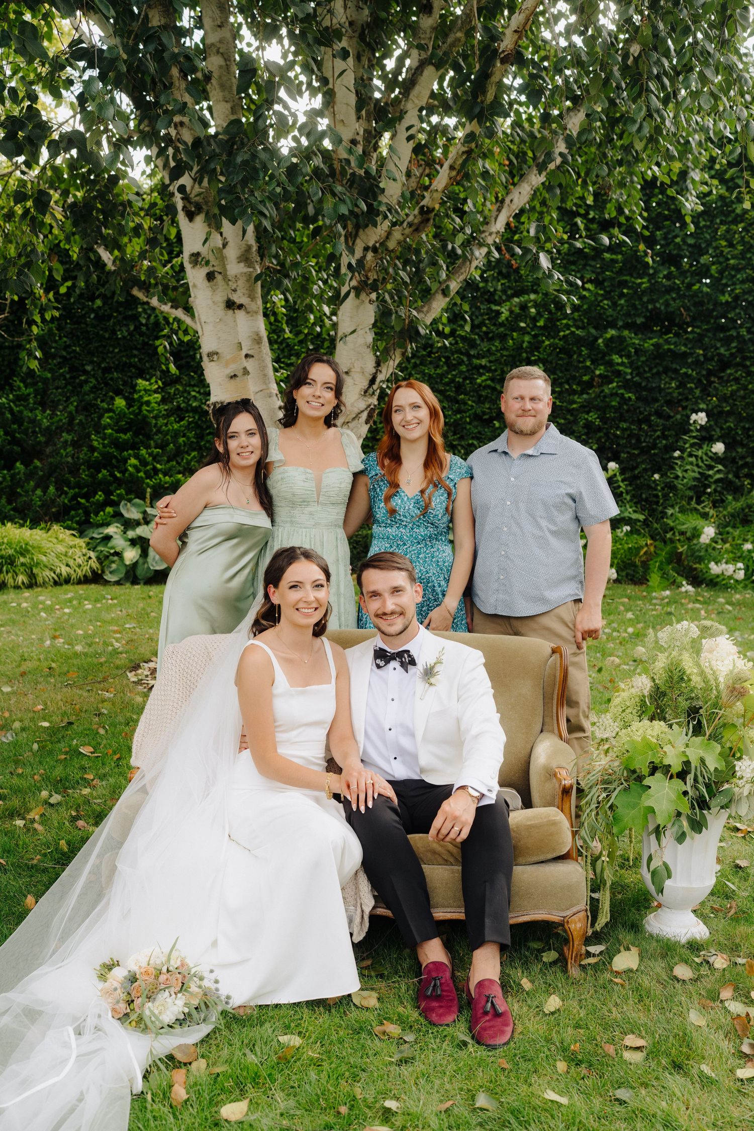 Wedding party lounges on vintage couch in lush garden setting with white floral decorations.