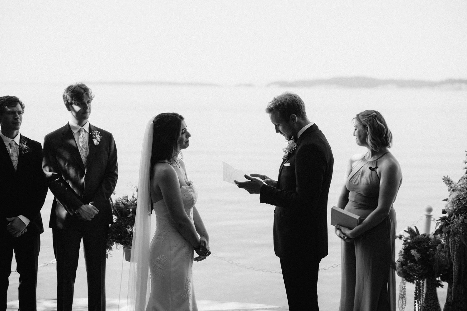 Black and white photo of a wedding ceremony with attendants gathered on a deck overlooking the water.
