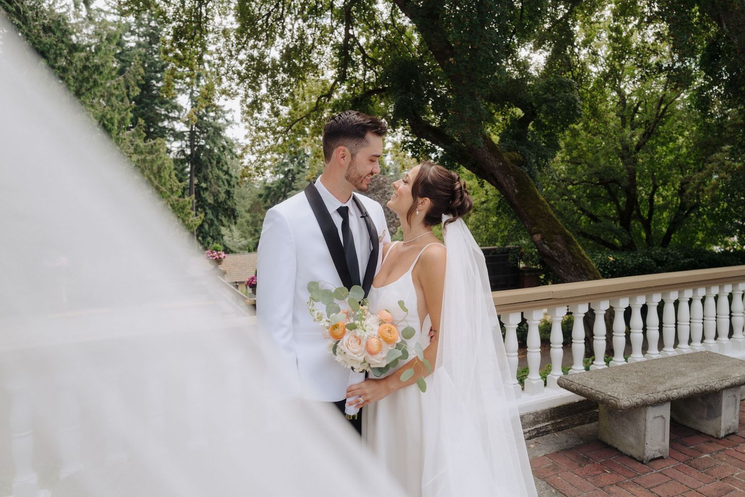 A couple shares an intimate moment on a terrace with white balustrades and lush trees.