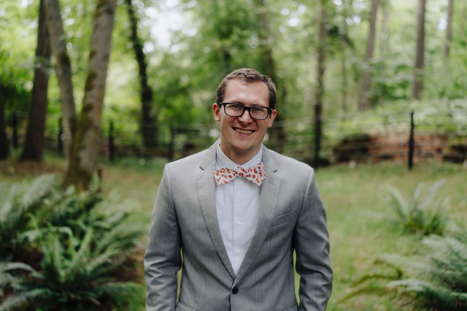 Groomsmen in grey suits and bow ties stand among ferns and evergreen trees at an outdoor wedding.