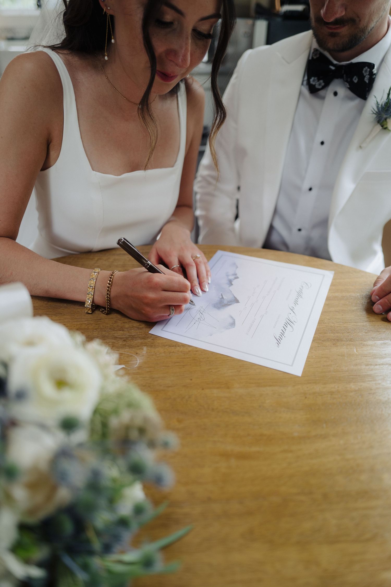 Close-up of hands signing wedding documents on a wooden table with floral decorations.