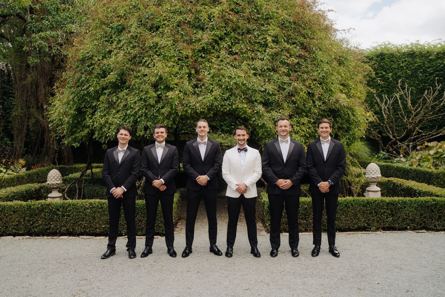Groomsmen in formal attire pose for photos in front of garden hedges.