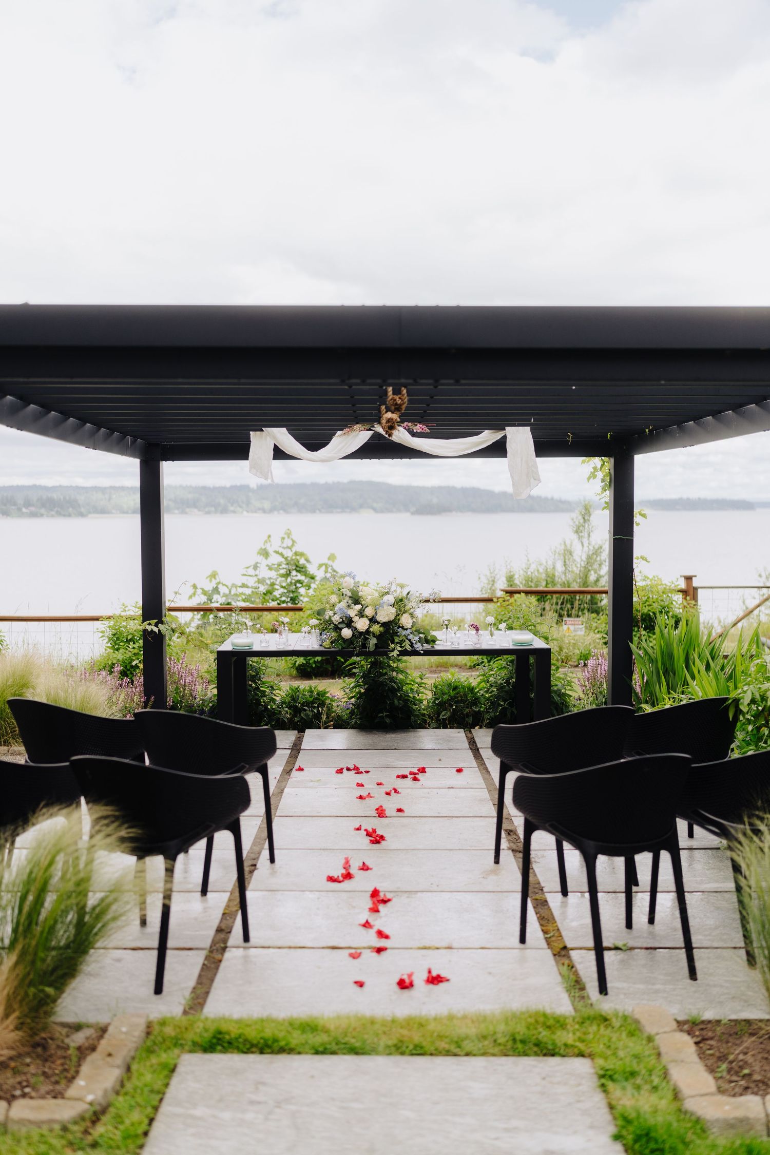 Modern black chairs line an outdoor deck with scattered red rose petals and water views.
