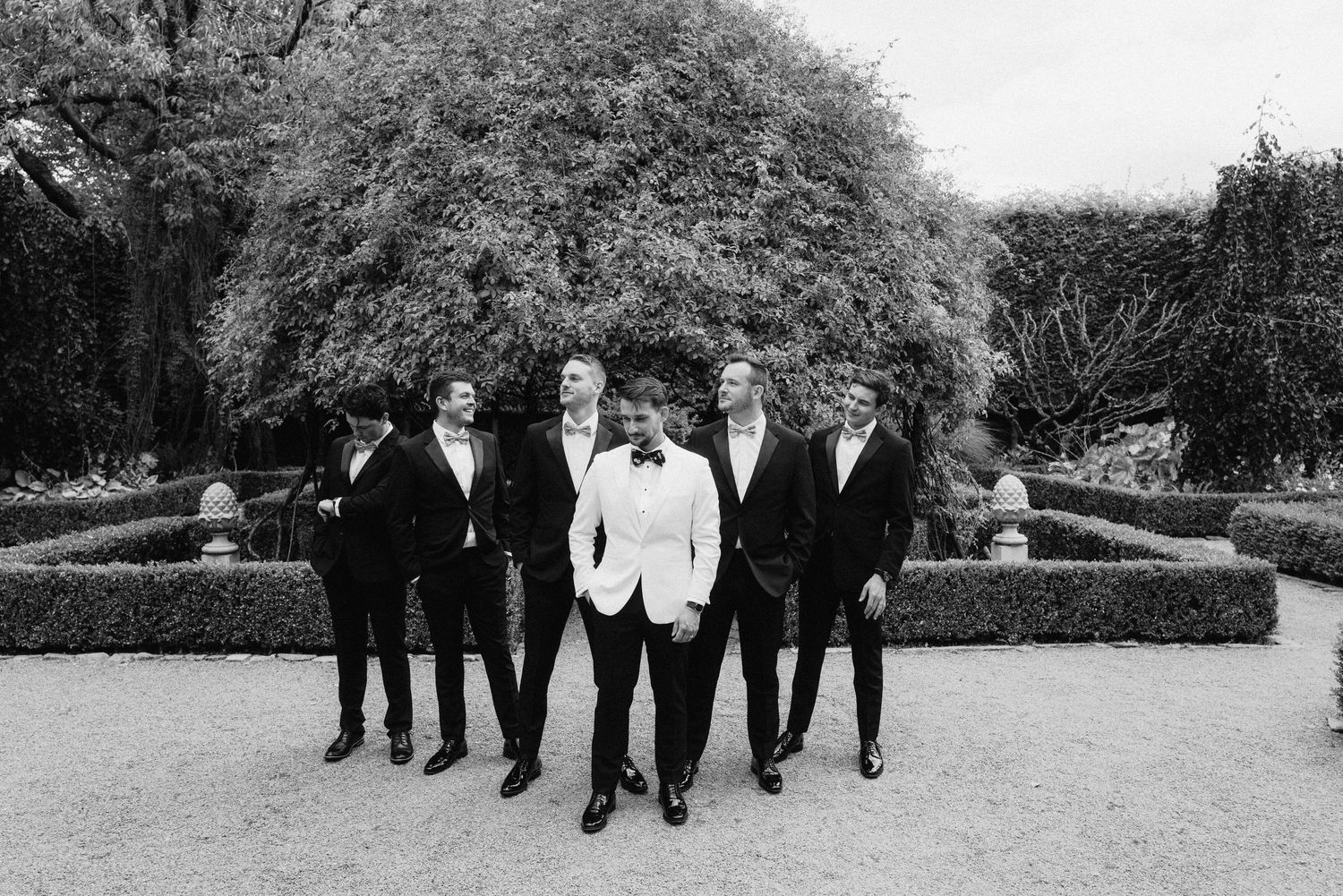 Group in formal attire poses in front of manicured garden hedges in black and white.