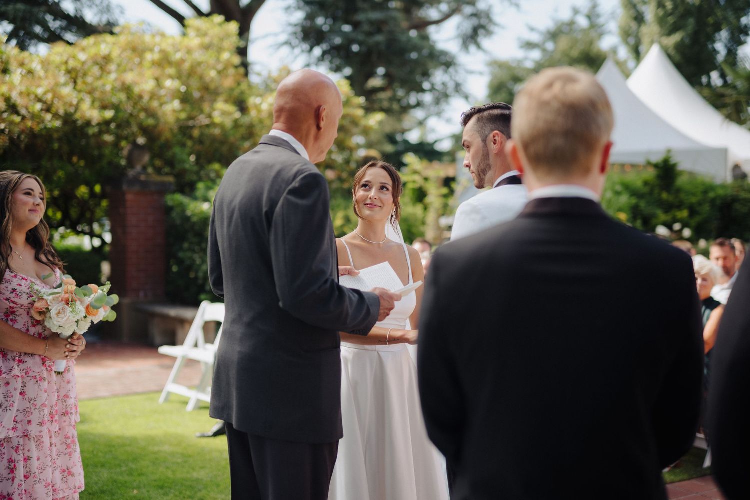 Wedding ceremony guests gather outdoors under trees during a summer garden celebration.