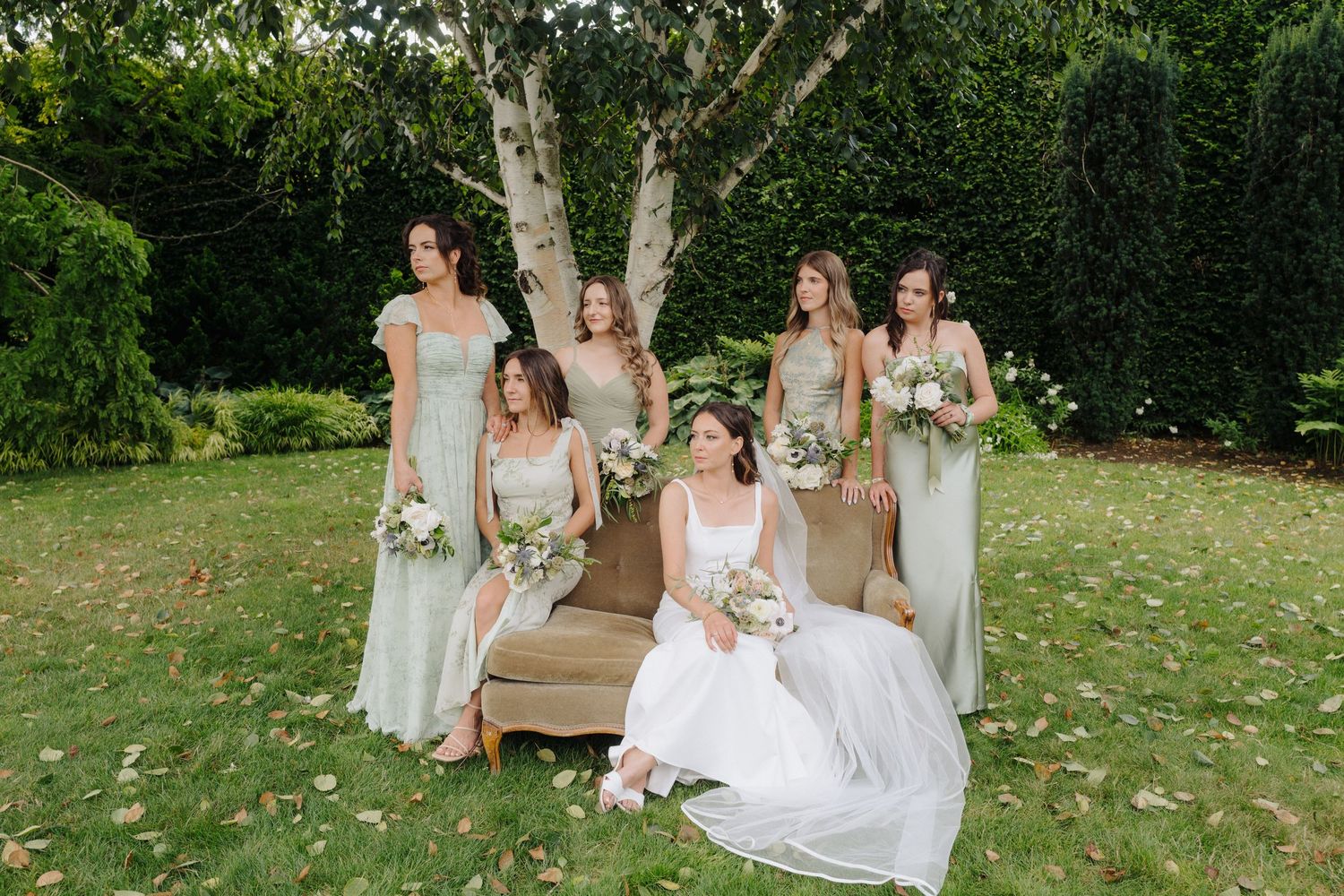 Wedding party poses on rustic wooden bench in garden setting with bridesmaids in sage green dresses.