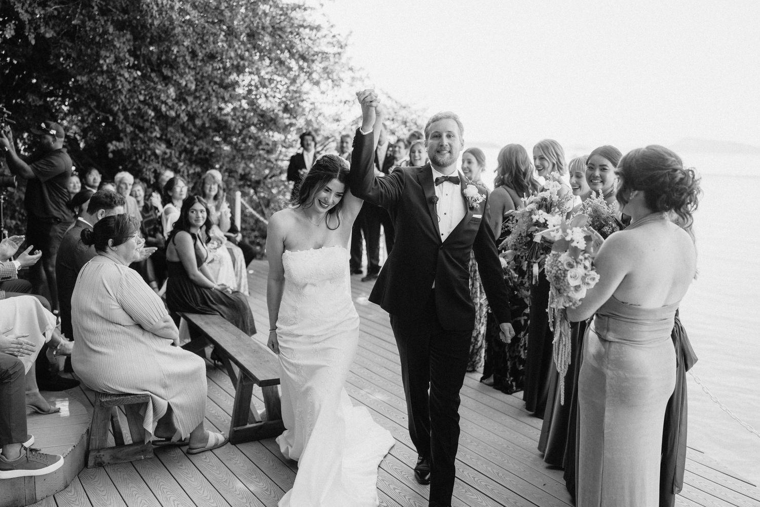 Black and white photo of wedding guests watching an outdoor waterfront ceremony.