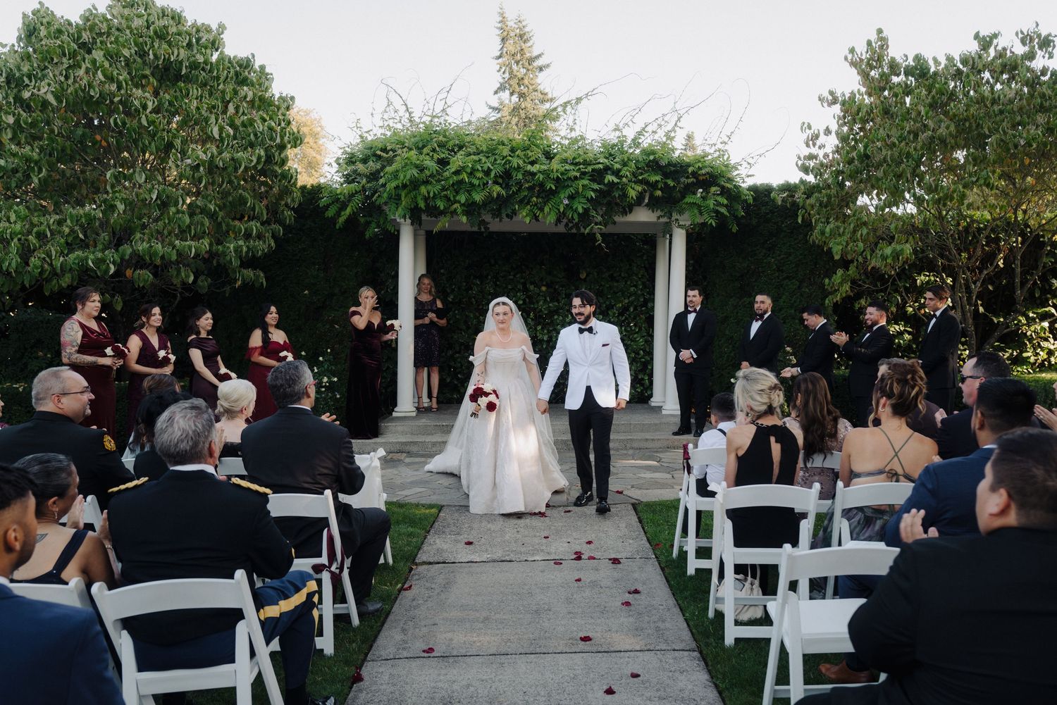 An outdoor wedding ceremony setup with white chairs and guests gathering under trees at dusk.