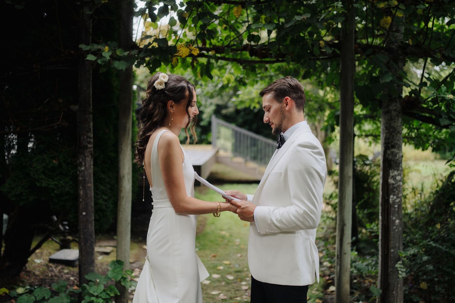 A couple shares an intimate moment during their outdoor wedding ceremony beneath a canopy of green trees.